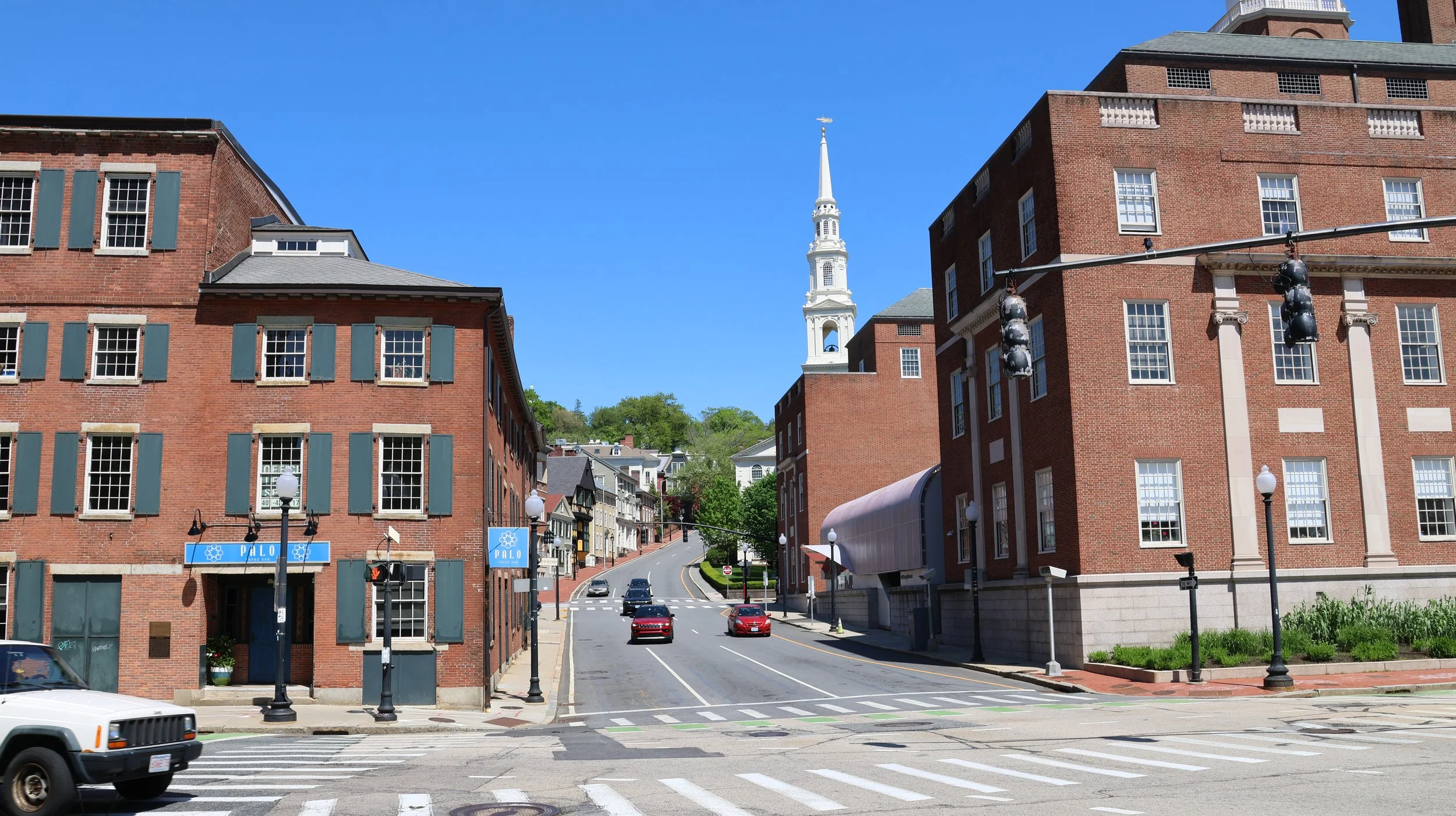 A city street scene with brick buildings, a church steeple in the background, a traffic light, cars on the road, and street lamps under a clear blue sky.