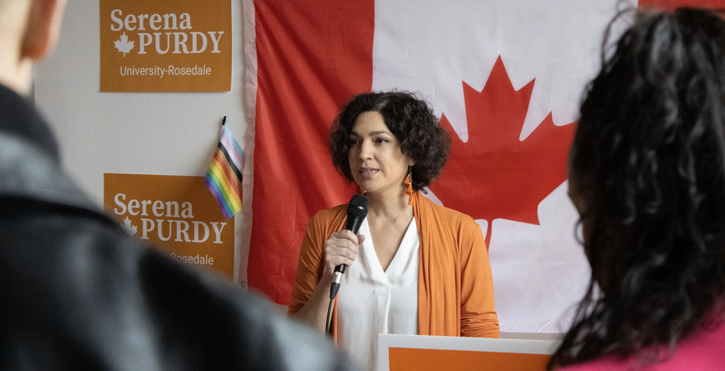 A woman standing in front of a Canadian flag and a Serena Purdy campaign sign, speaking into a microphone, with people in the audience listening.