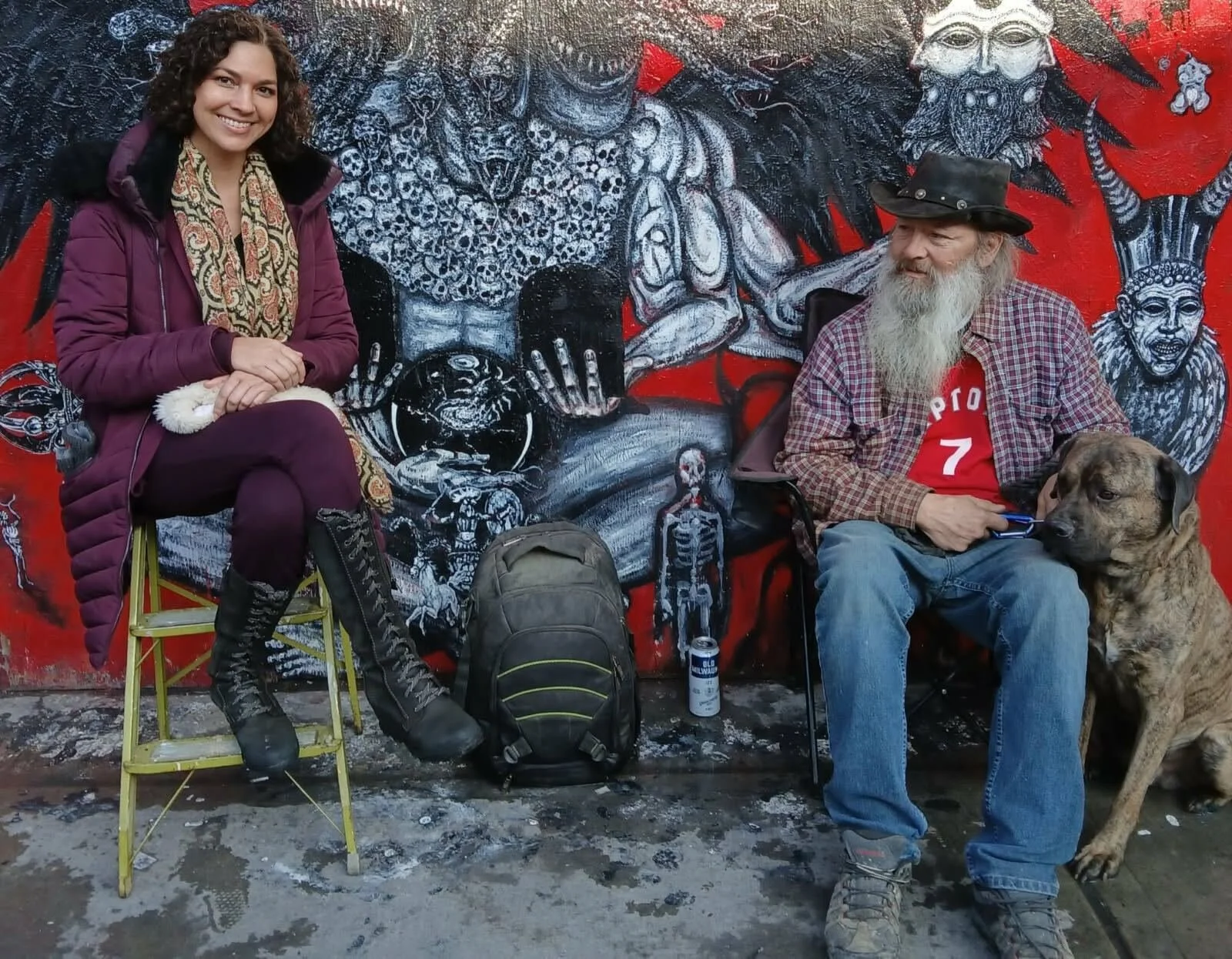 A woman with curly dark hair smiling, sitting on a yellow step stool, wearing a purple coat and patterned scarf, alongside an elderly man with a beard, wearing a black hat and checkered shirt, sitting with a large dog, in front of a colorful mural featuring abstract faces and figures.