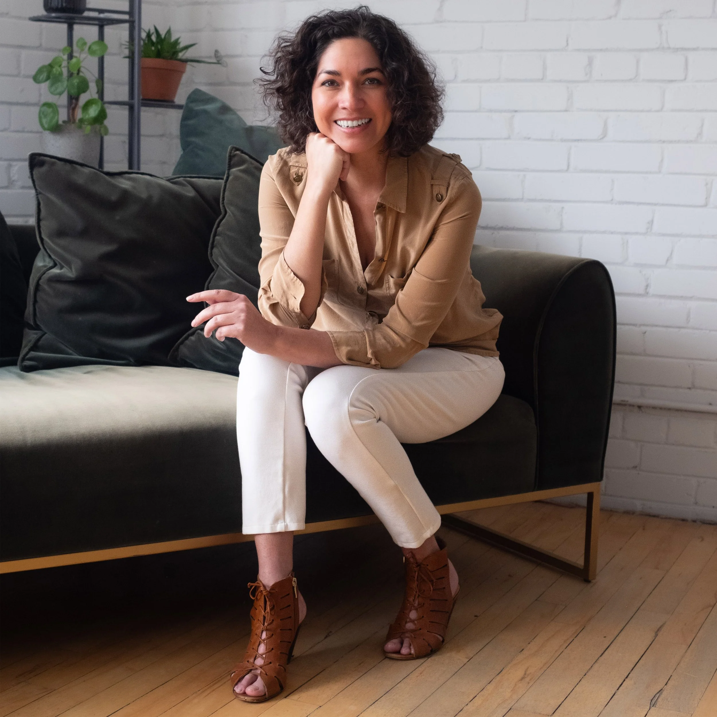 A woman with curly dark hair sitting on a black sofa with beige pants and brown peep-toe heels, smiling in front of a white brick wall with plants on the shelf behind her.