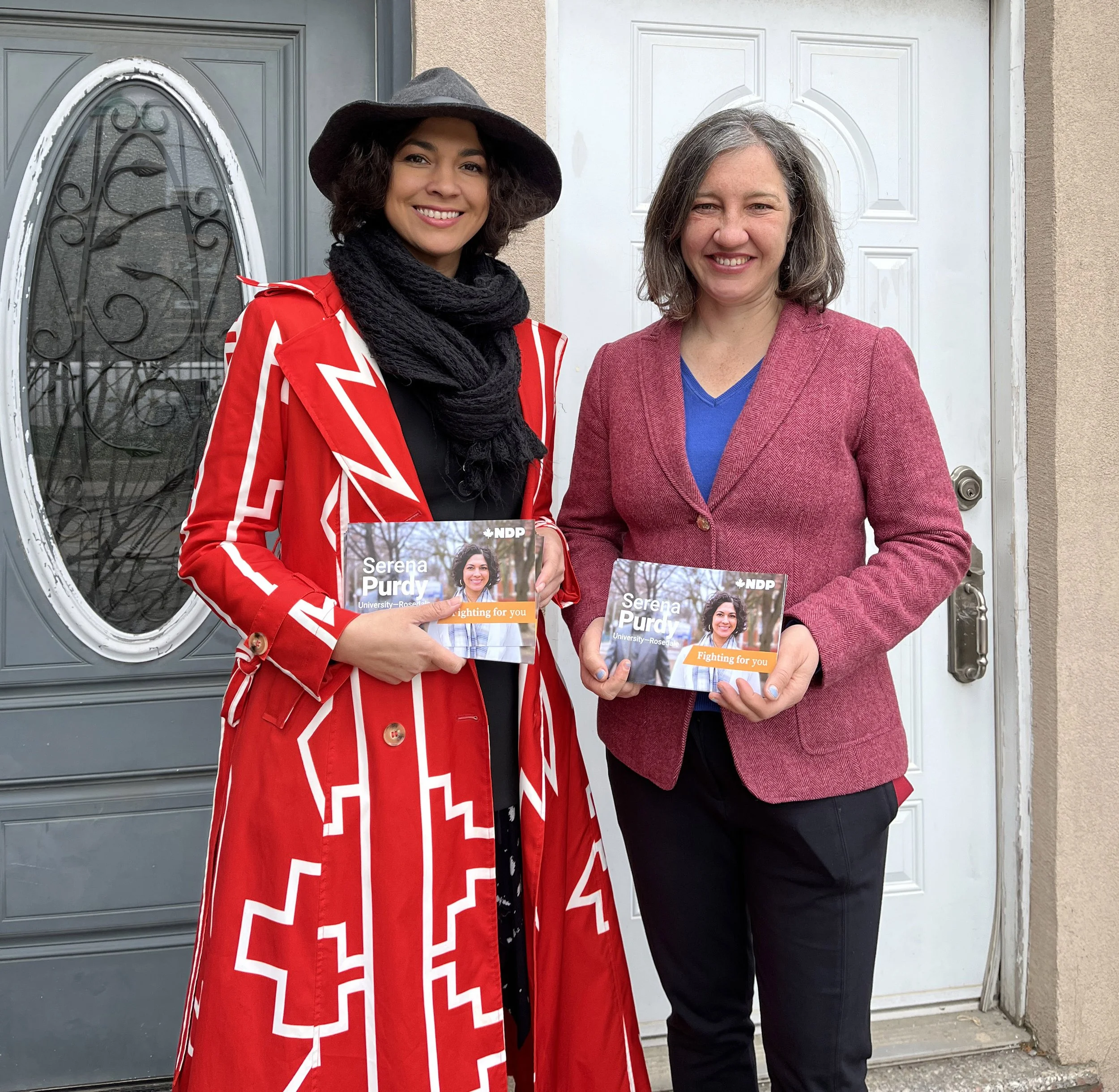 Two women standing in front of a house door holding campaign flyers for Serena Purdy, both smiling and dressed in colorful clothing.