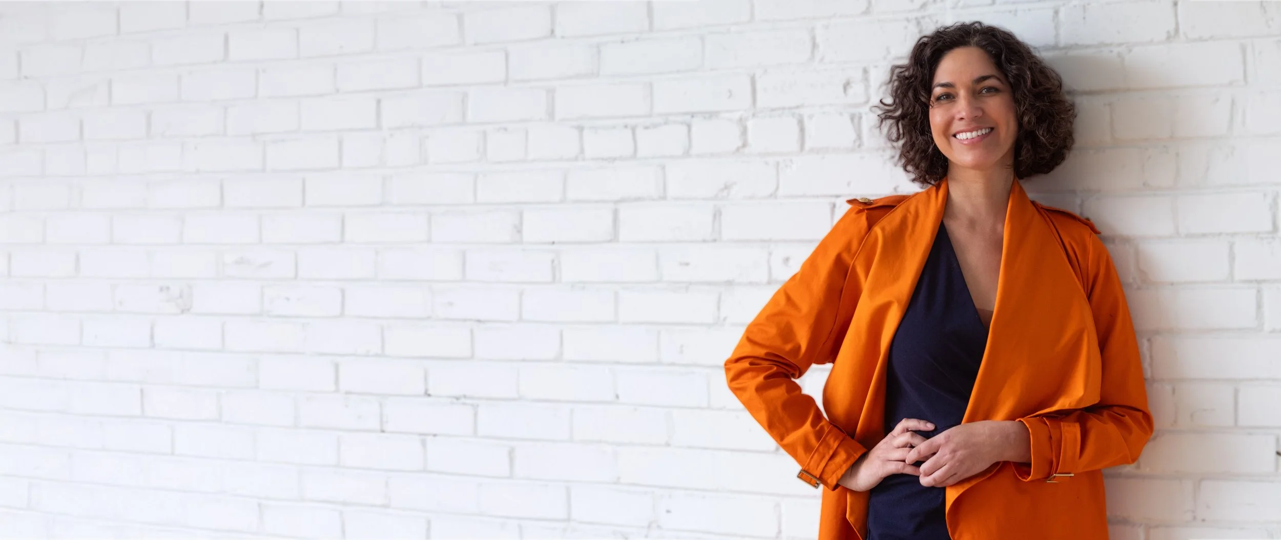 Smiling woman with curly hair wearing an orange jacket and black top, standing against a white brick wall.