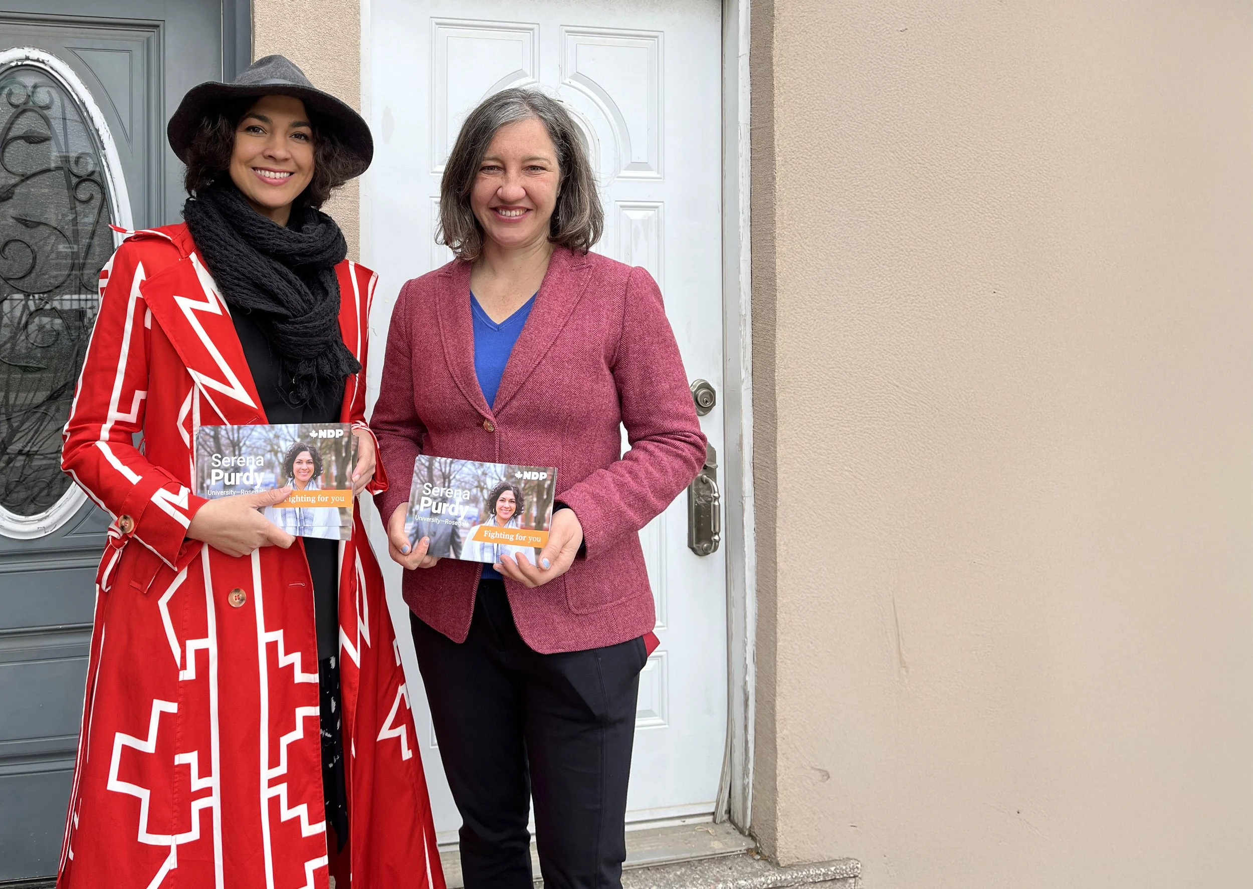 Two women stand outside, holding political campaign flyers featuring Serena Purdy. The woman on the left wears a wide-brimmed hat, black scarf, and red and white jacket with bold patterns. The woman on the right wears a pink blazer over a blue shirt. They are smiling and standing in front of a door and a plain beige wall.
