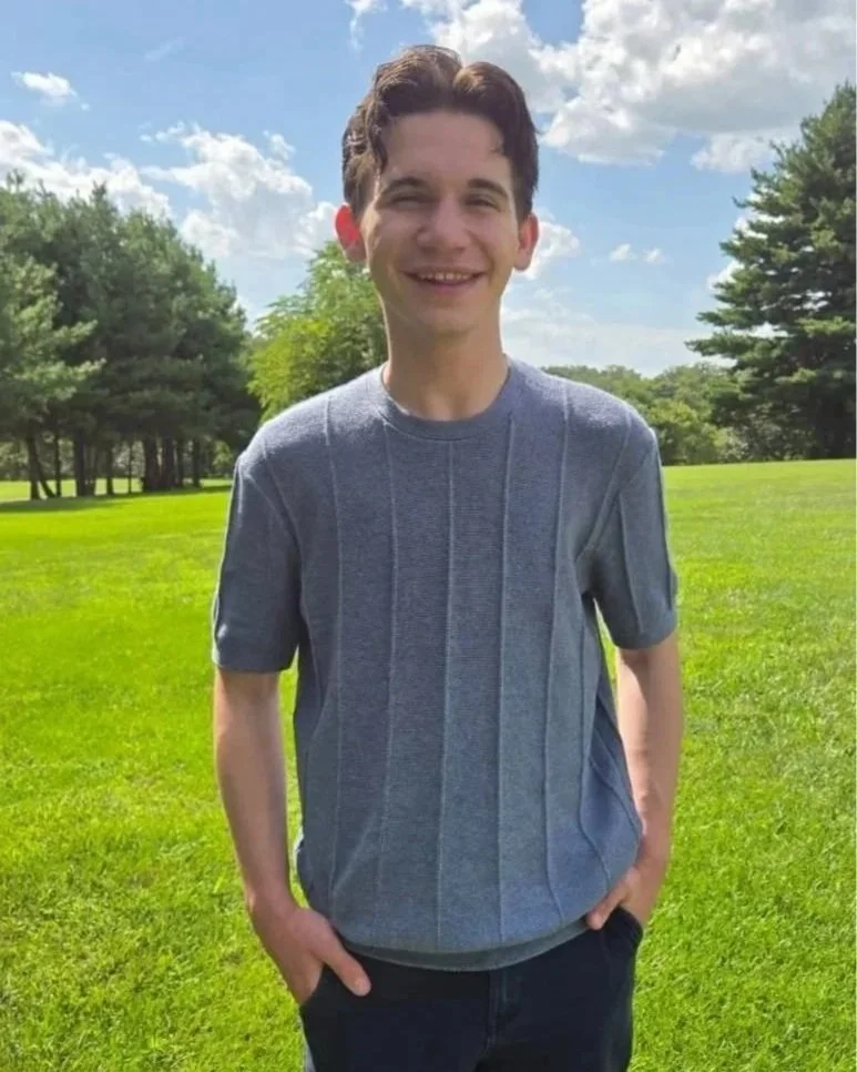 Young man smiling outdoors in a park, wearing a gray t-shirt, with green grass and trees in the background under a partly cloudy sky.