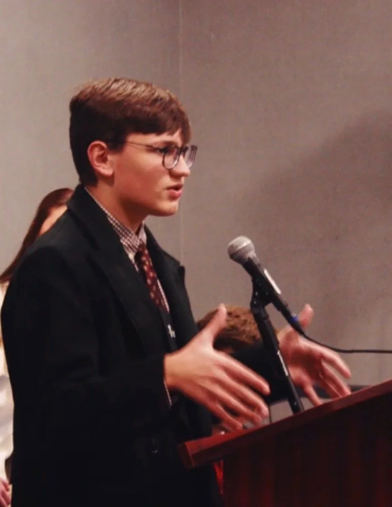 A young man standing at a podium gives an impassioned address in a dark black suit with a polka dot tie.