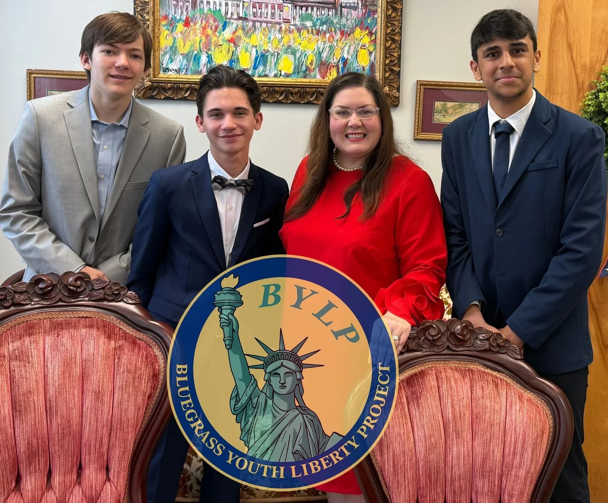 A group of four people, three young men and one wom, standing together at a formal event. They are dressed in suits and a red dress, with a background featuring colorful artwork and framed pictures. The woman is holding a circular sign with the Statue of Liberty and the words " and "YOUTH LIBERTY." They are smiling and posing for the photo.
