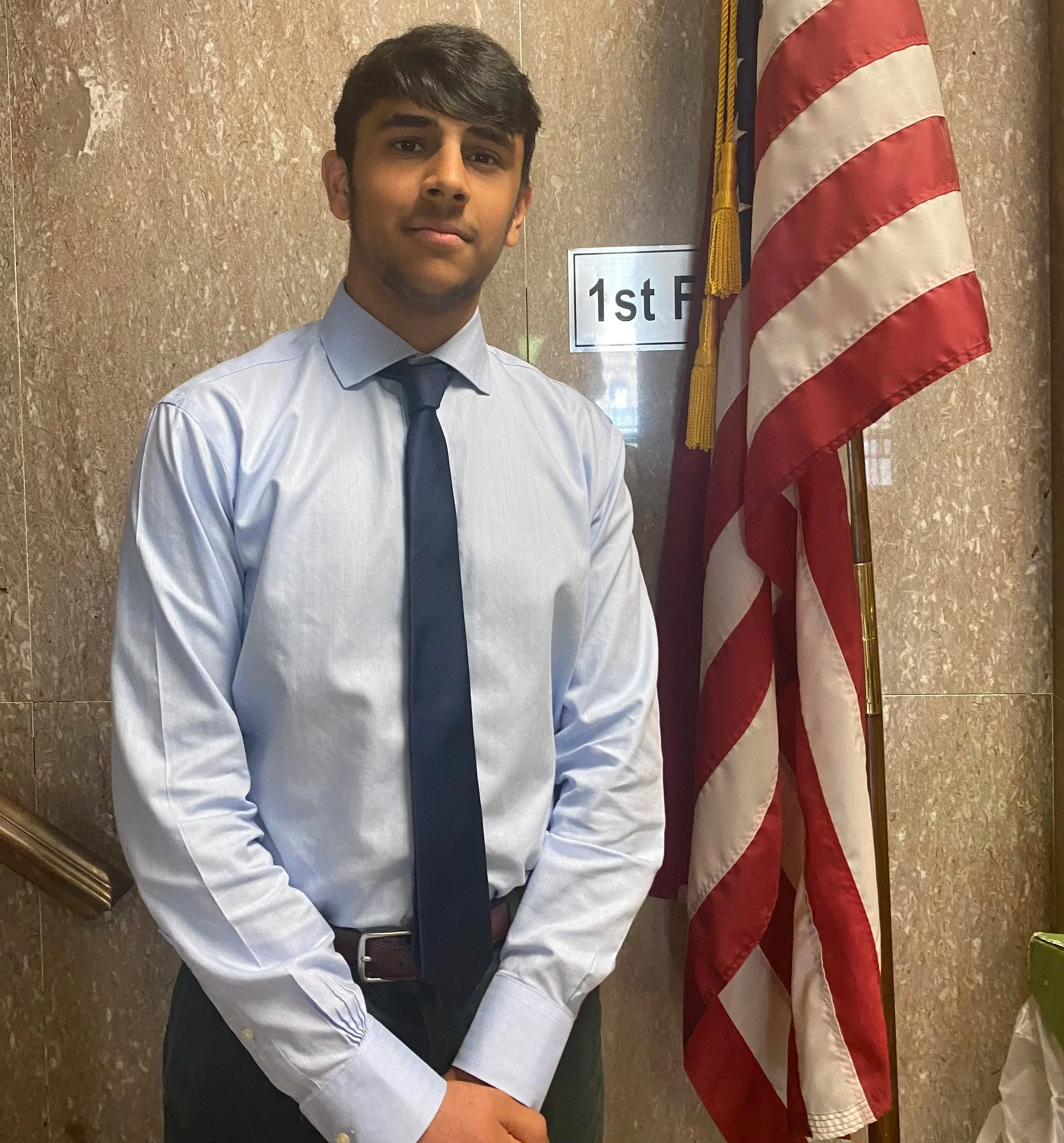 A young man dressed in a light blue shirt and dark tie, standing in front of an American flag and a sign that reads '1st F'. He is posing for the photo with a neutral expression on a beige marble wall background.