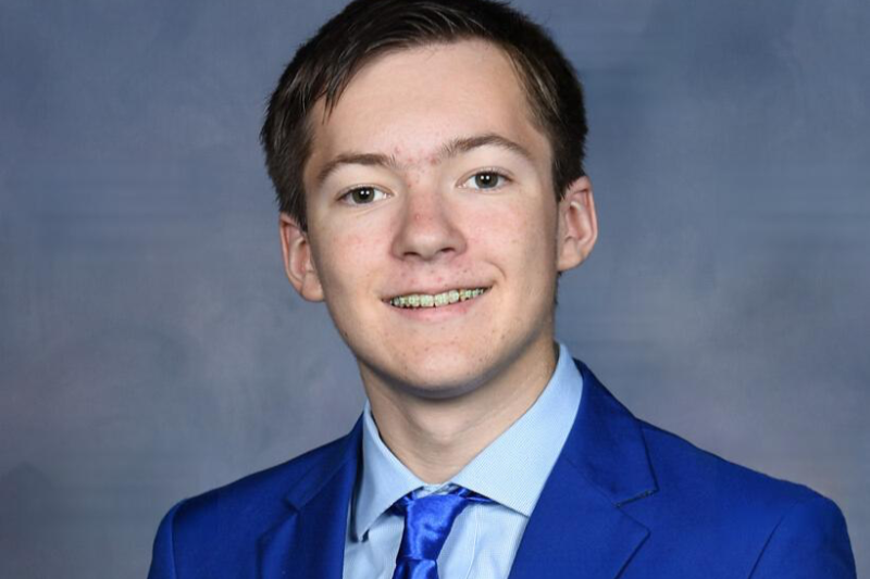 A young man with short brown hair, wearing a blue suit, light blue shirt, and blue tie, smiling at the camera against a gray background.