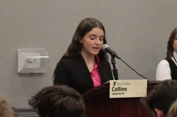 Young girl speaking at a podium with a microphone, wearing a black blazer and pink shirt, in a formal setting.
