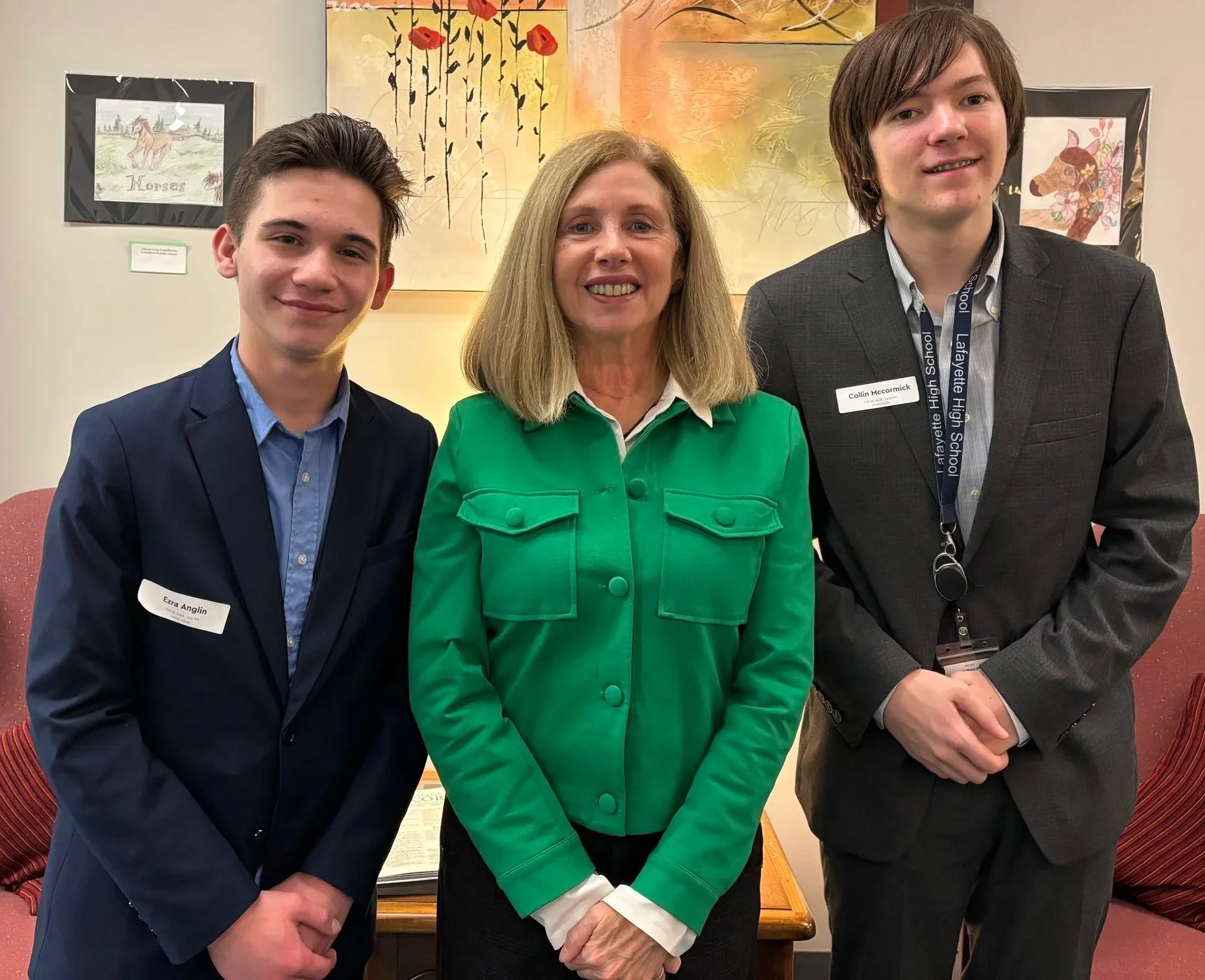 Three people standing side by side indoors, smiling at the camera. The woman in the middle has blonde hair and is wearing a green jacket. The young man on the left has dark hair and is dressed in a blue shirt and dark blazer. The young man on the right has brown hair and wears a gray suit with a lanyard and a name tag. There are colorful paintings on the wall behind them.