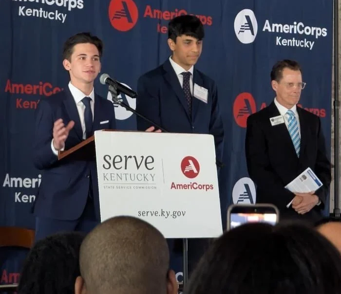 Three men in suits at a podium during a ceremony or event, with a backdrop displaying logos for AmeriCorps and Kentucky, and a sign reading 'serve KENTUCKY' with agency logos.