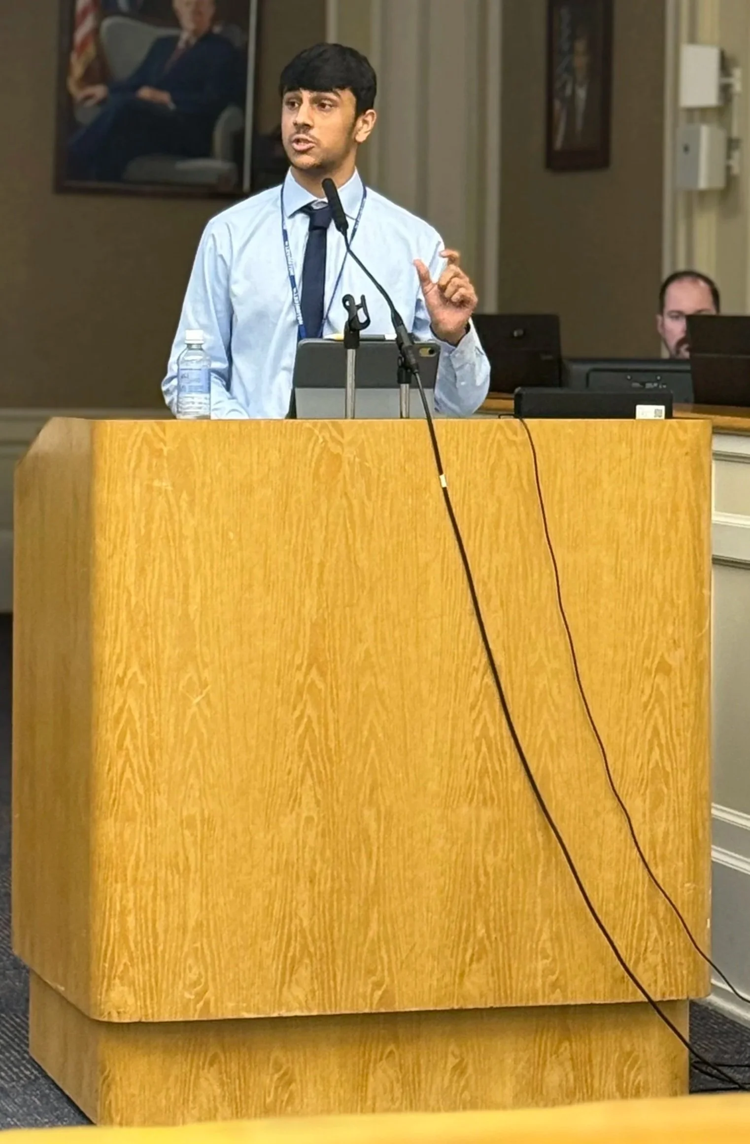 A man with black hair, wearing a light blue shirt and a dark blue tie, stands behind a wooden podium speaking into a microphone. There is a water bottle on the podium. In the background, a person is sitting at a computer, and a large framed portrait of a person wearing a dark suit is hanging on the wall.