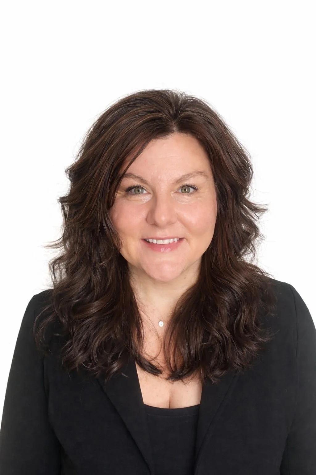 A woman with shoulder-length wavy brown hair wearing a black blazer and a black top, smiling against a plain white background.