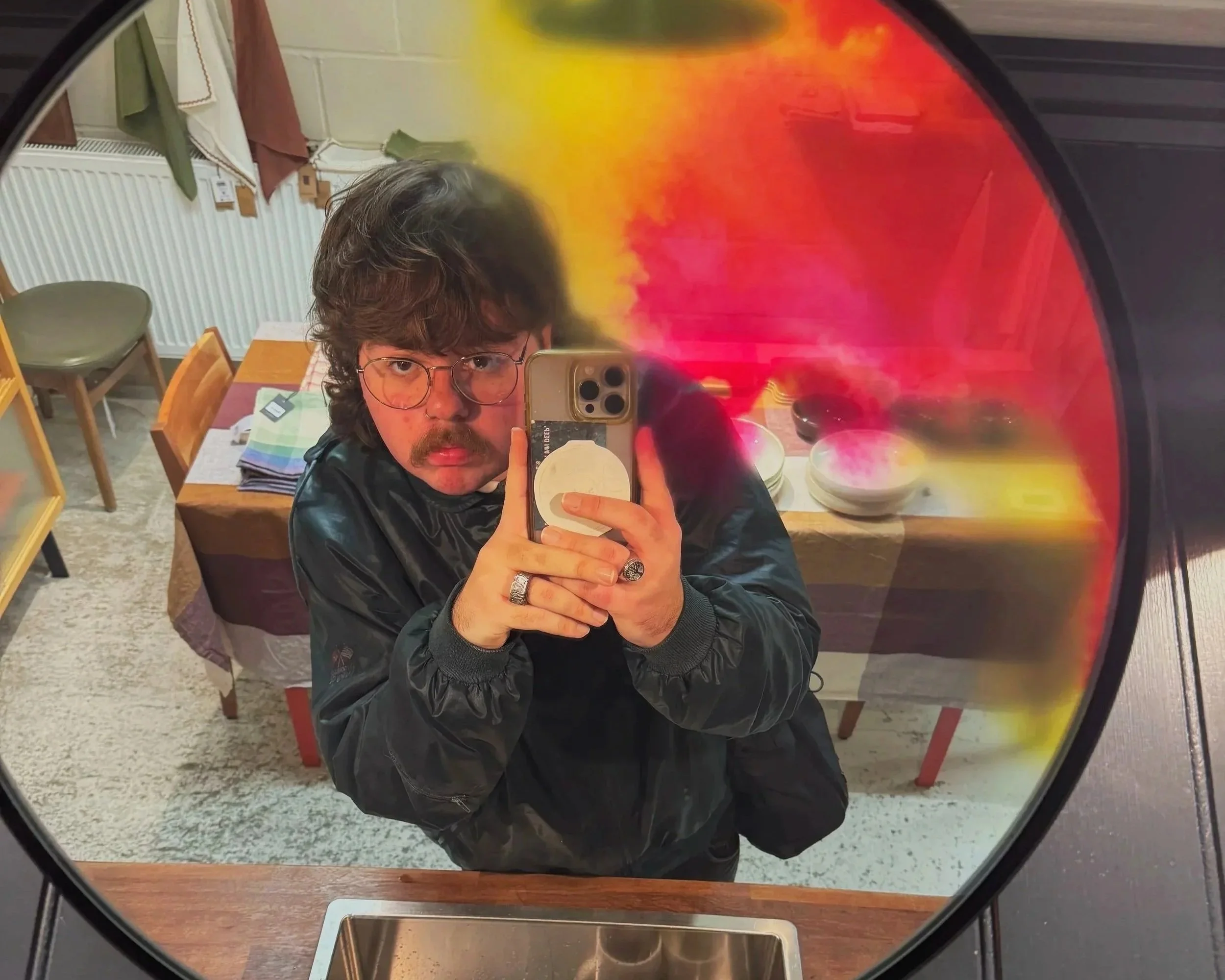 A person with glasses and curly hair taking a selfie in a mirror that reflects a colorful abstract painting, a dining room with chairs, and a person sitting at a table.