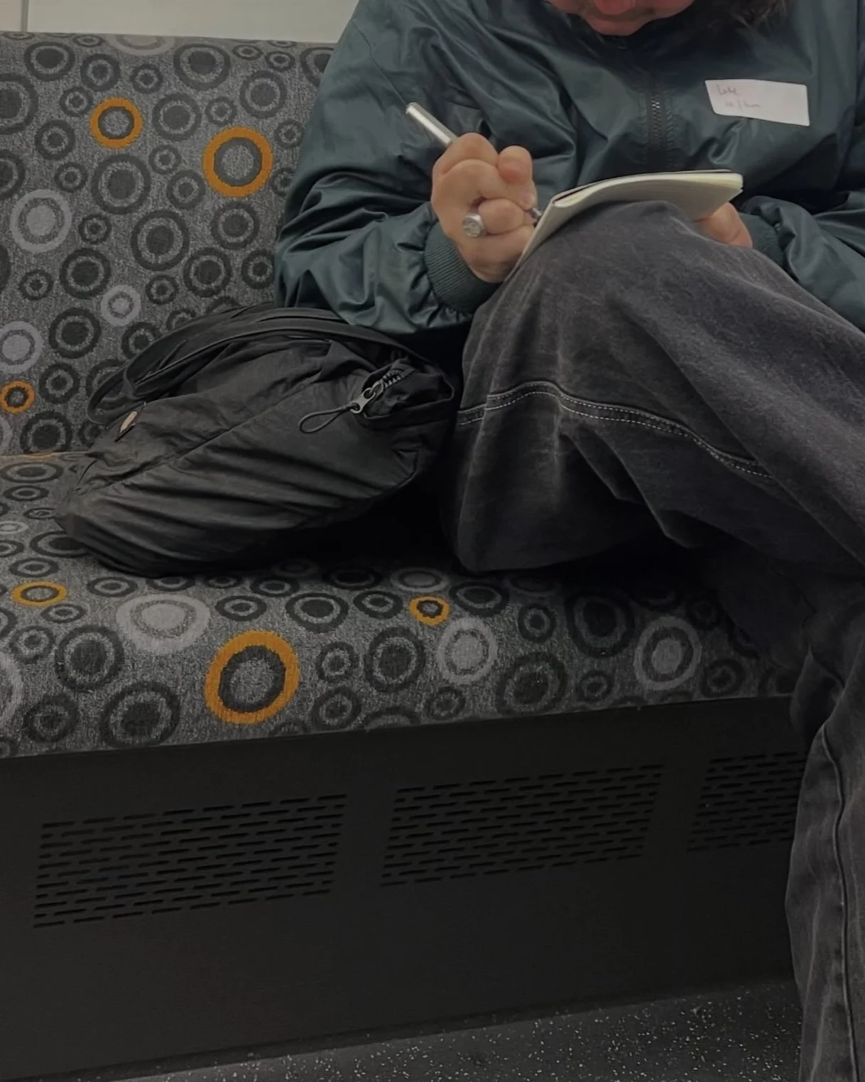 Person sitting on a patterned fabric bench, writing in a small notebook with a pen. They wear a dark gray jacket and dark jeans. A black backpack is placed next to them on the bench.