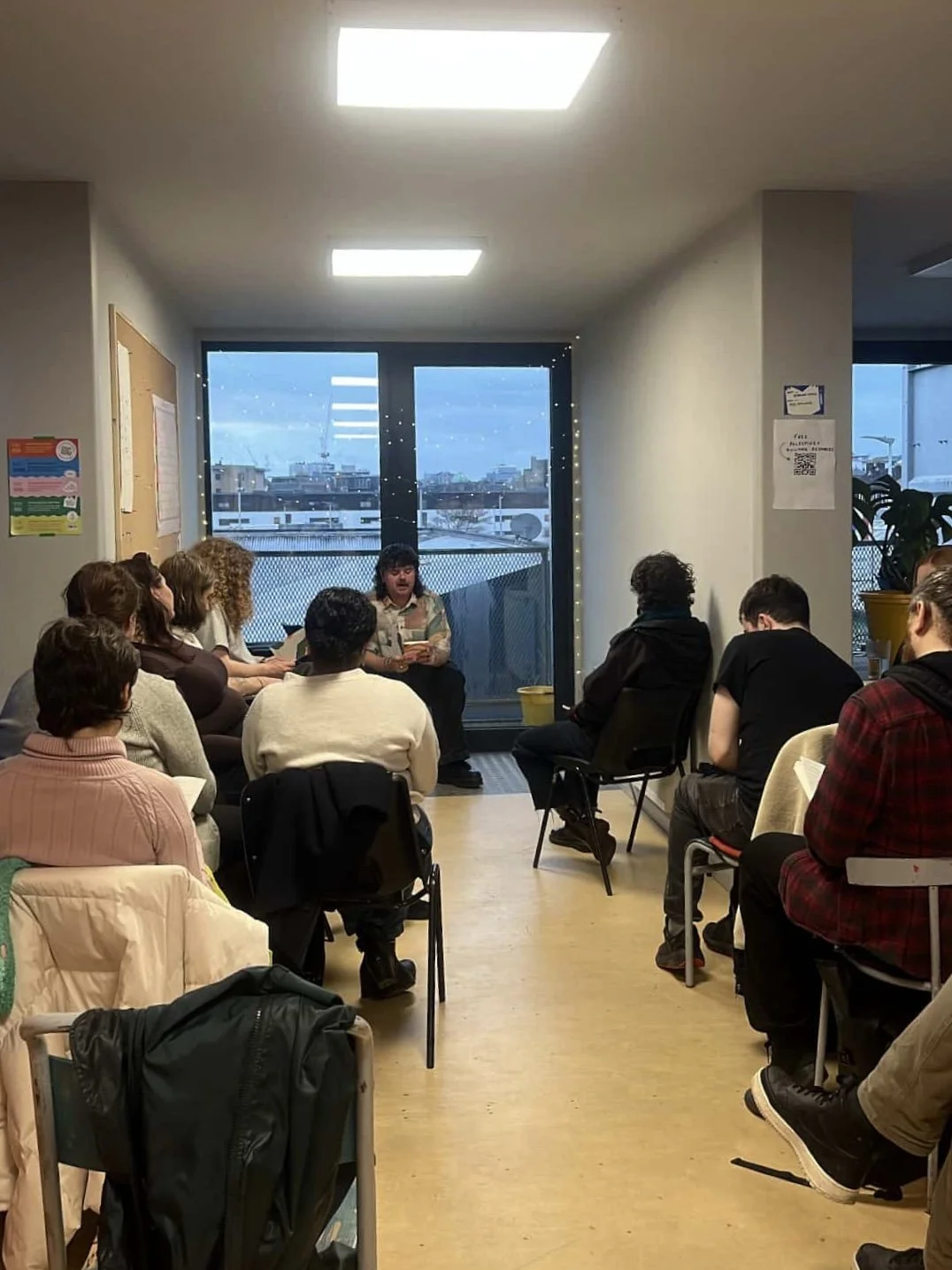 A small group of people sitting in chairs in a circle in a room, listening to a woman speaking. The room has large windows with cityscape views and string lights.