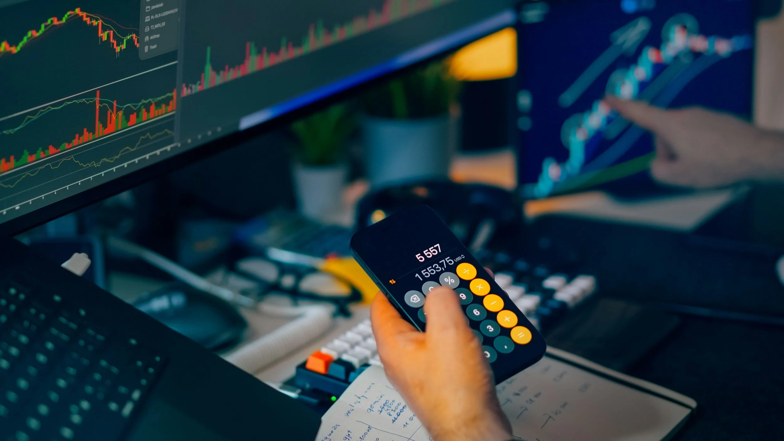 Close-up of a person using a calculator on a cluttered desk with multiple computer screens displaying stock market charts in the background.