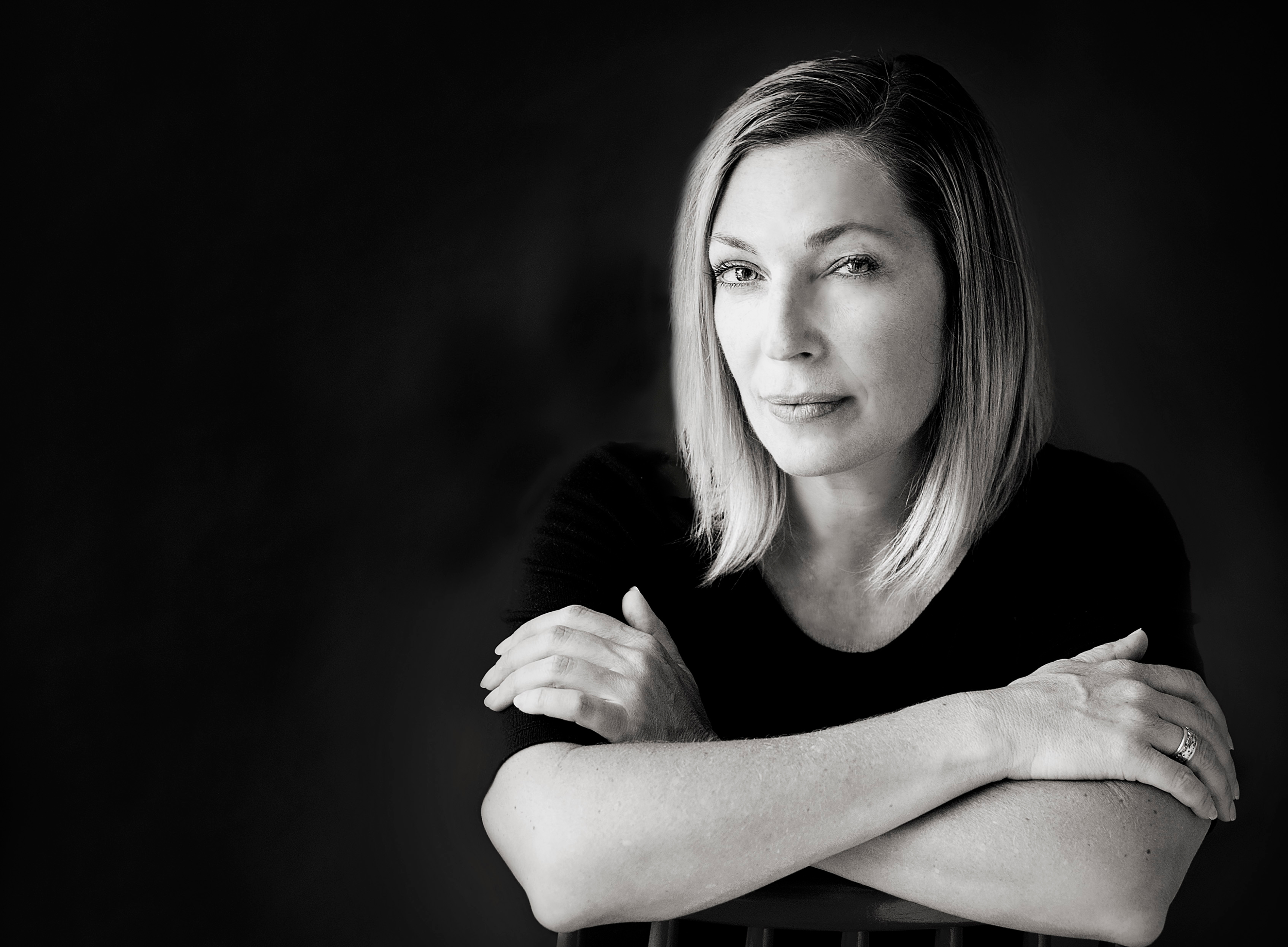 Black and white portrait of a woman with shoulder-length hair, wearing a black top, with her arms crossed on a chair, looking at the camera.