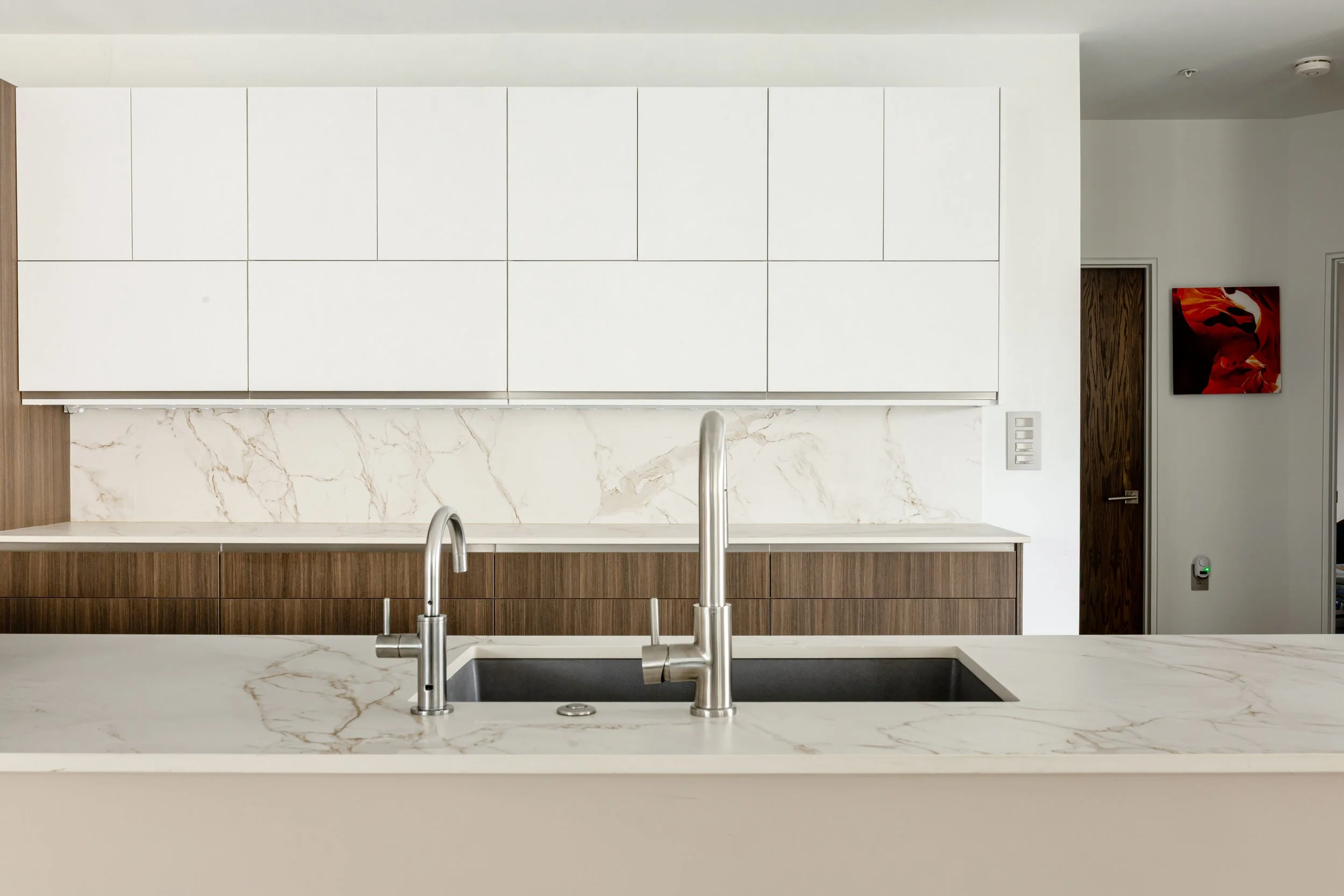 Modern kitchen with white upper cabinets, wooden lower cabinets, marble backsplash, and a marble countertop with a stainless steel sink and faucet.