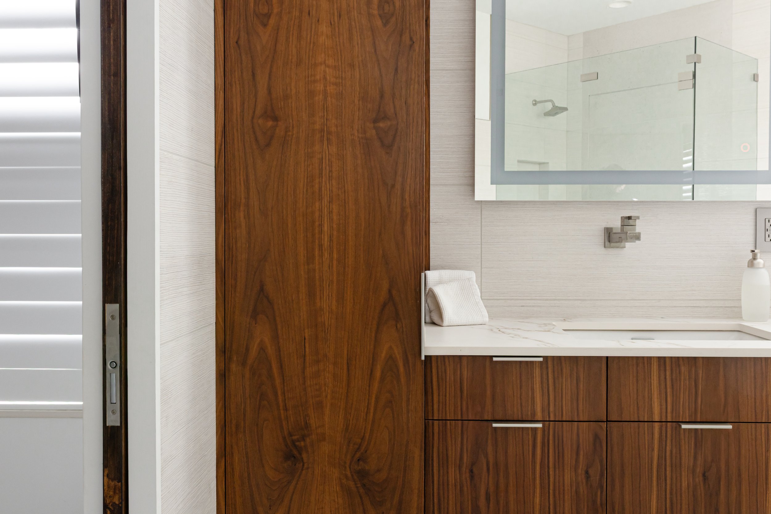 Modern bathroom with wooden cabinetry, white countertop, towel, mirror, and shower area reflected in the mirror.