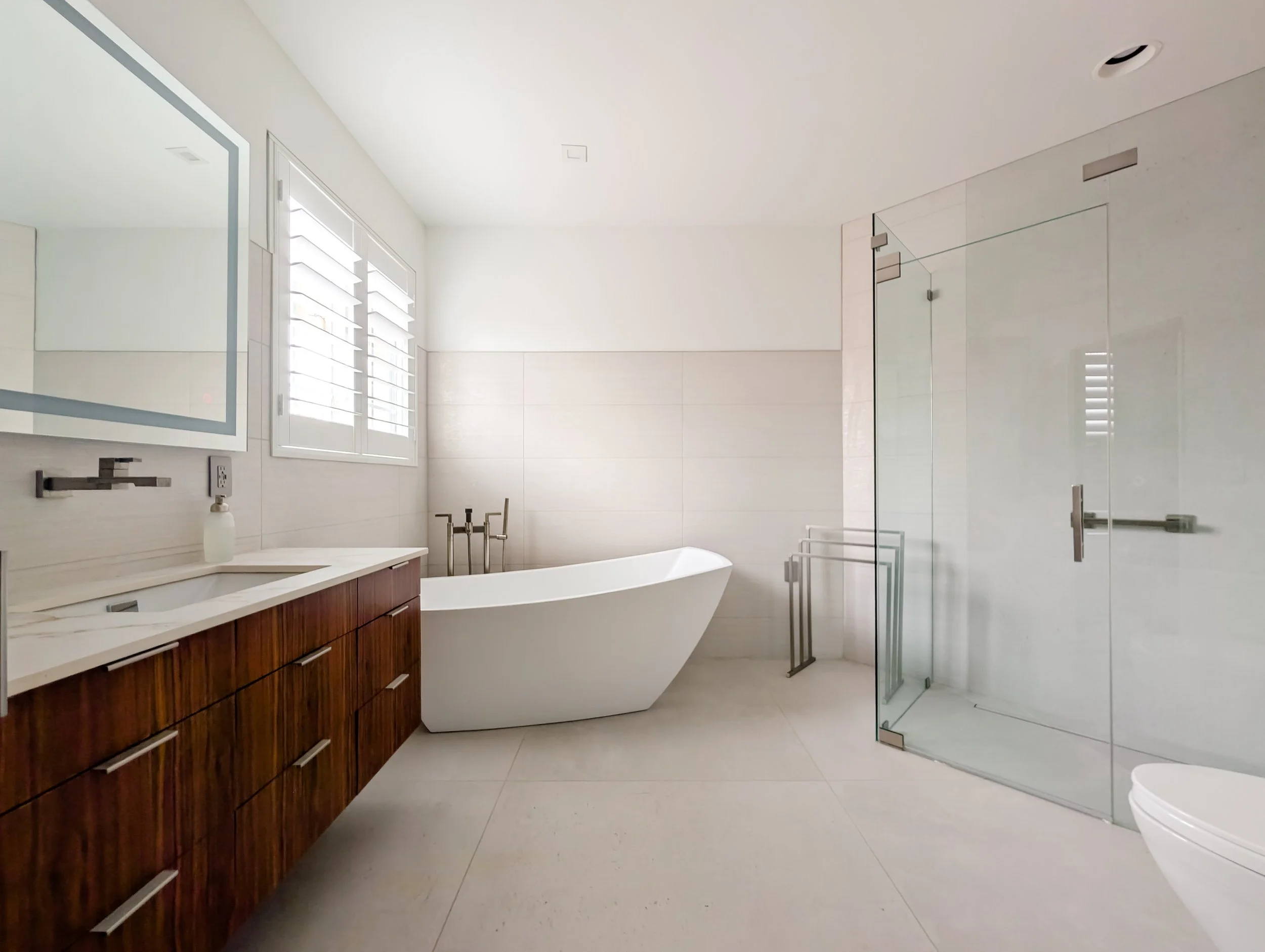 Modern bathroom with a wooden vanity, white sink, wall-mounted mirror, bathtub near the window, glass shower enclosure, and beige tiled floors and walls.