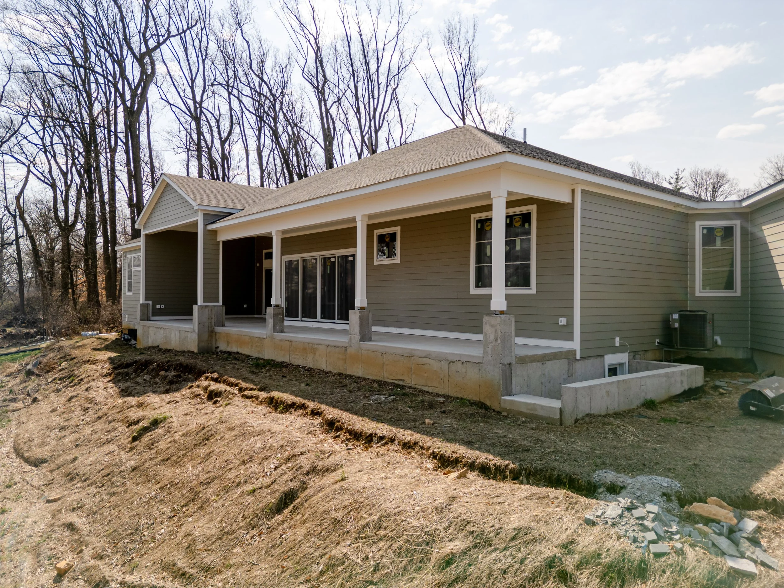 Newly constructed house with a covered porch, sliding glass doors, and windows, situated on a dirt yard with a wooded area in the background.