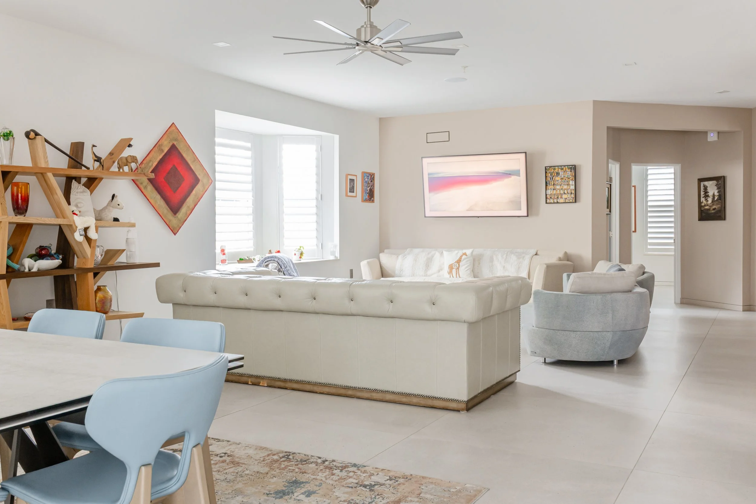 Bright, modern living room with white walls, white couch, light gray swivel chair, large window with white plantation shutters, decorative shelving with souvenirs and art, and abstract wall art.
