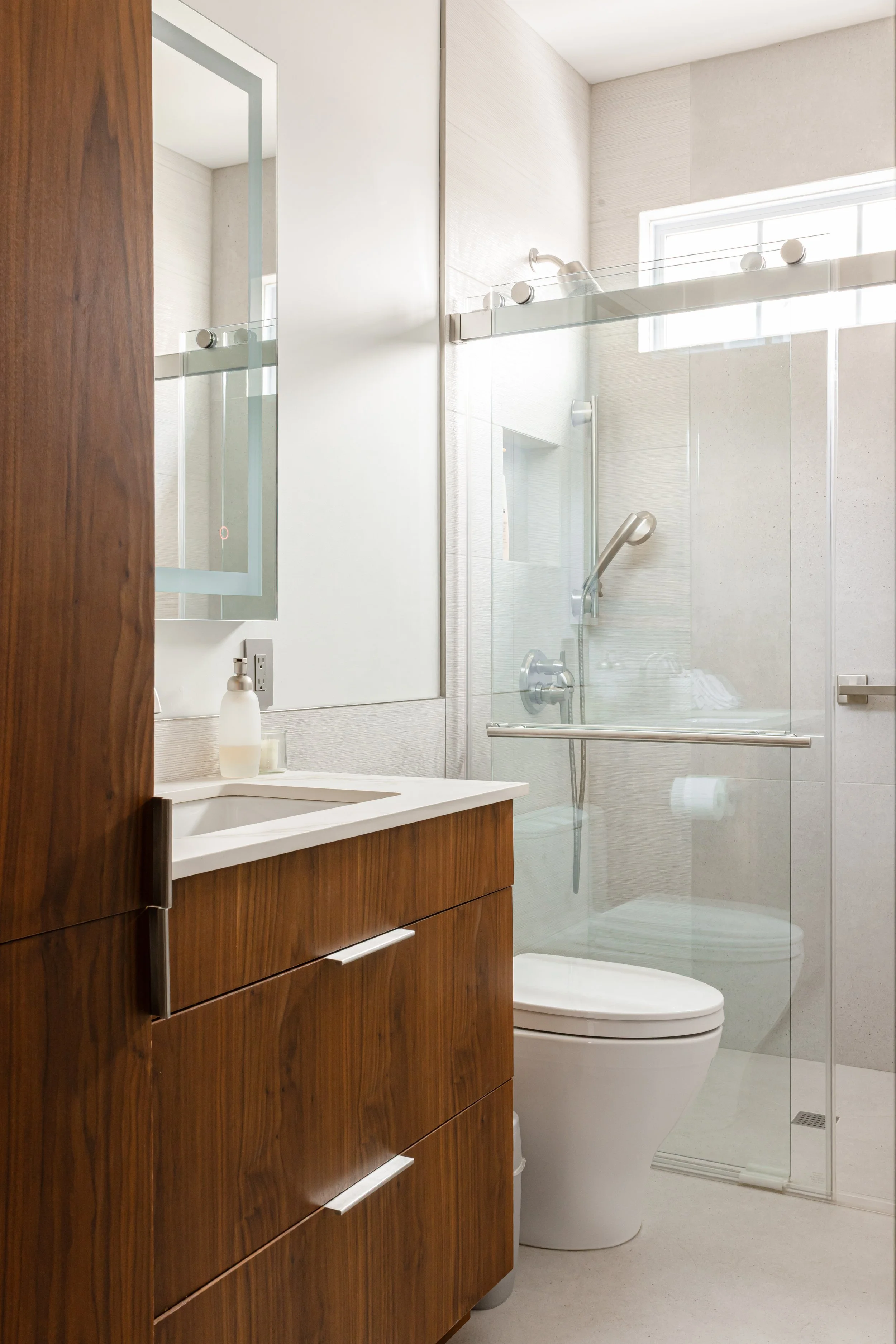 Modern bathroom with wooden vanity, white sink, soap dispenser, toilet, and glass-enclosed shower with natural light from a small window.