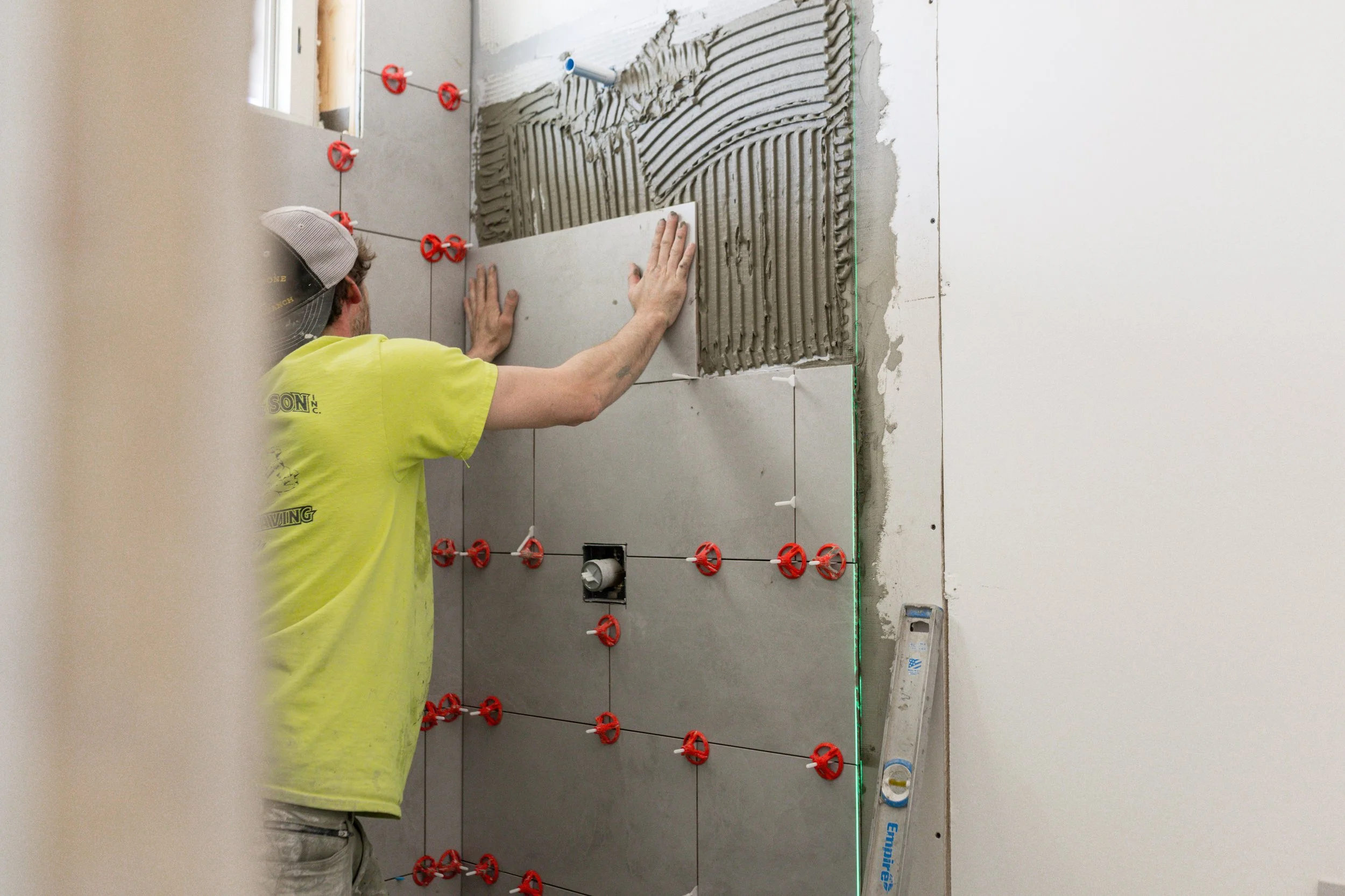 A construction worker installing large gray tiles on a wall, with a laser level marking the edge. The worker is wearing a yellow shirt and a gray cap, and is working on a partially finished bathroom or interior wall. Tiled section has round red space