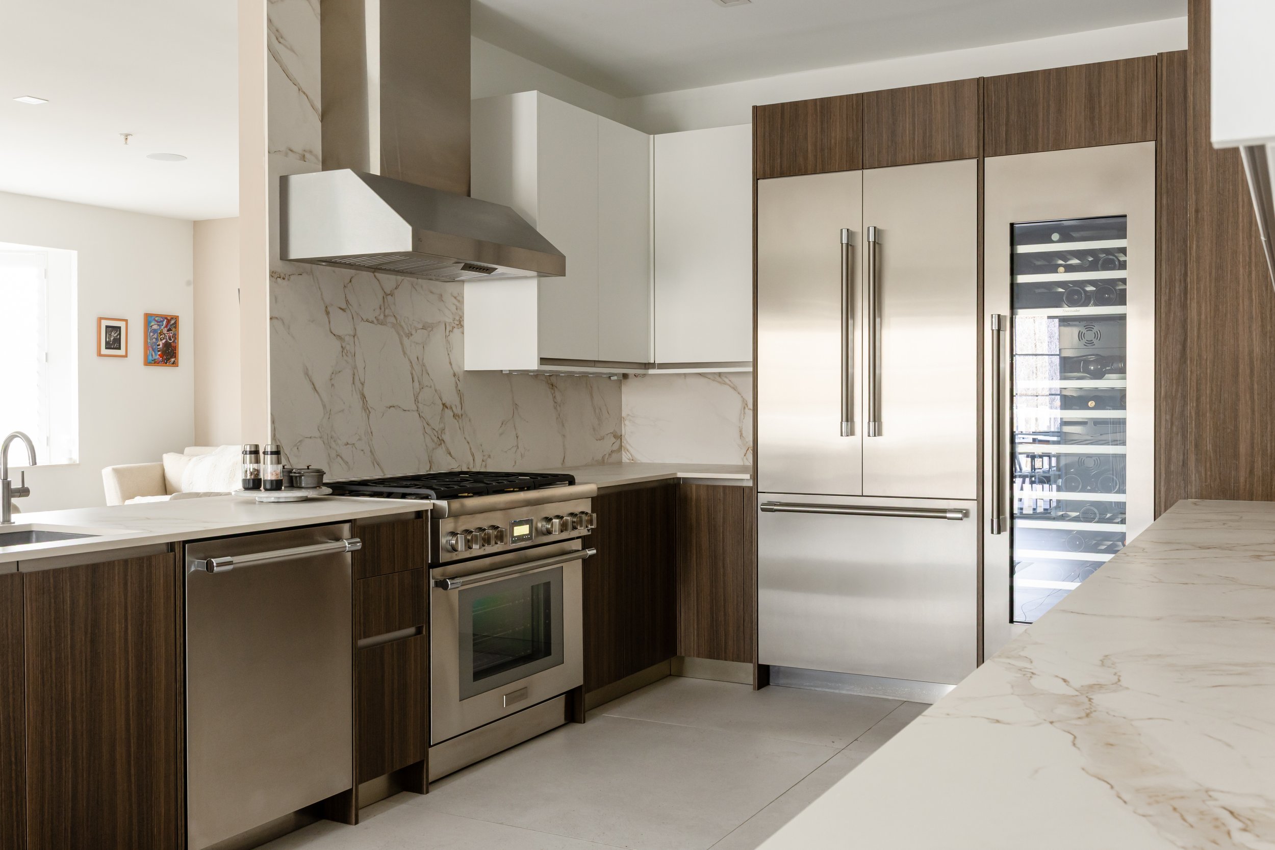Modern kitchen with stainless steel appliances, including a refrigerator, oven, and wine cooler, featuring wooden and white cabinetry, marble countertops, and a marble backsplash.