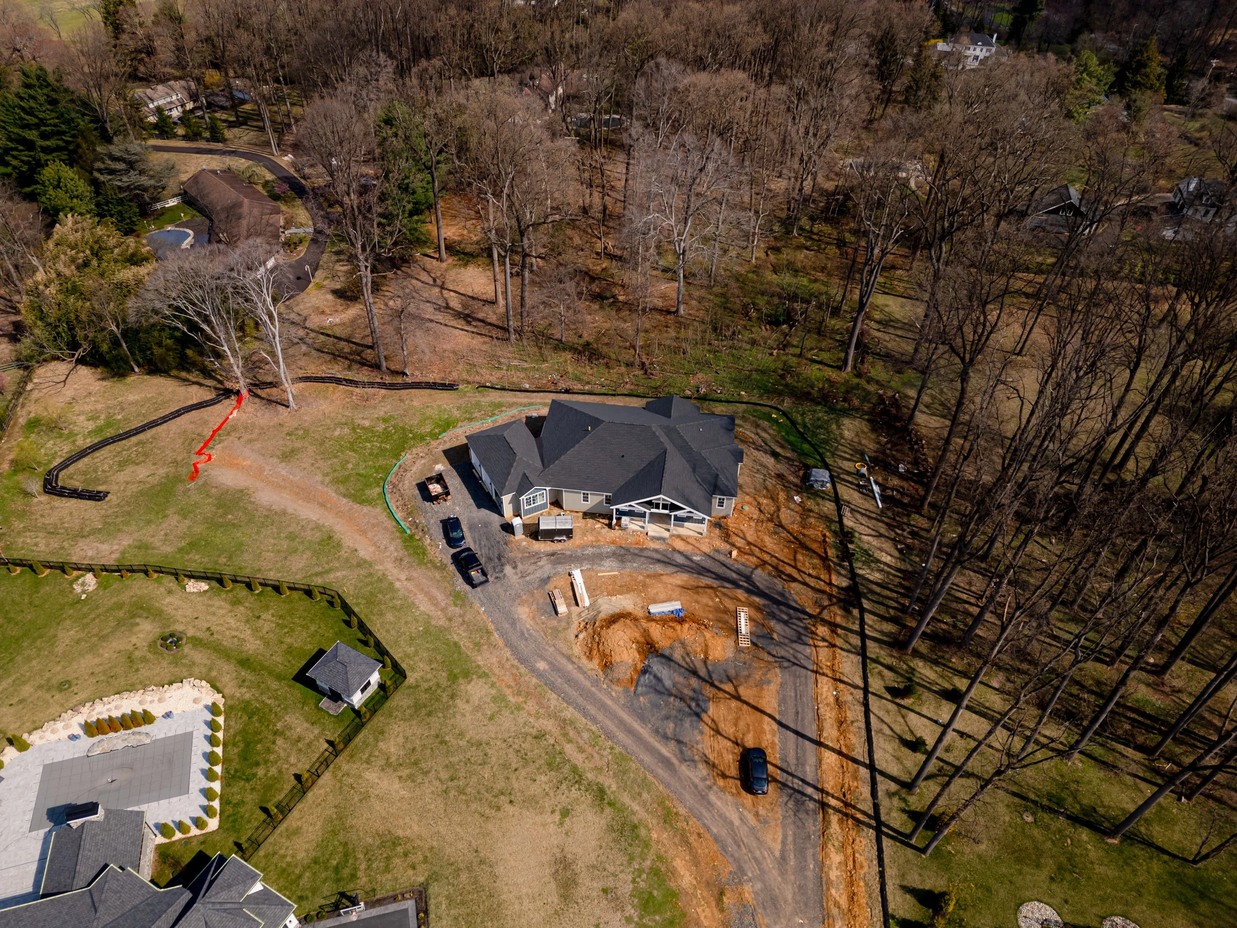 Aerial view of a house under construction, with dirt and gravel around it, surrounded by trees, neighboring houses, and a fence.