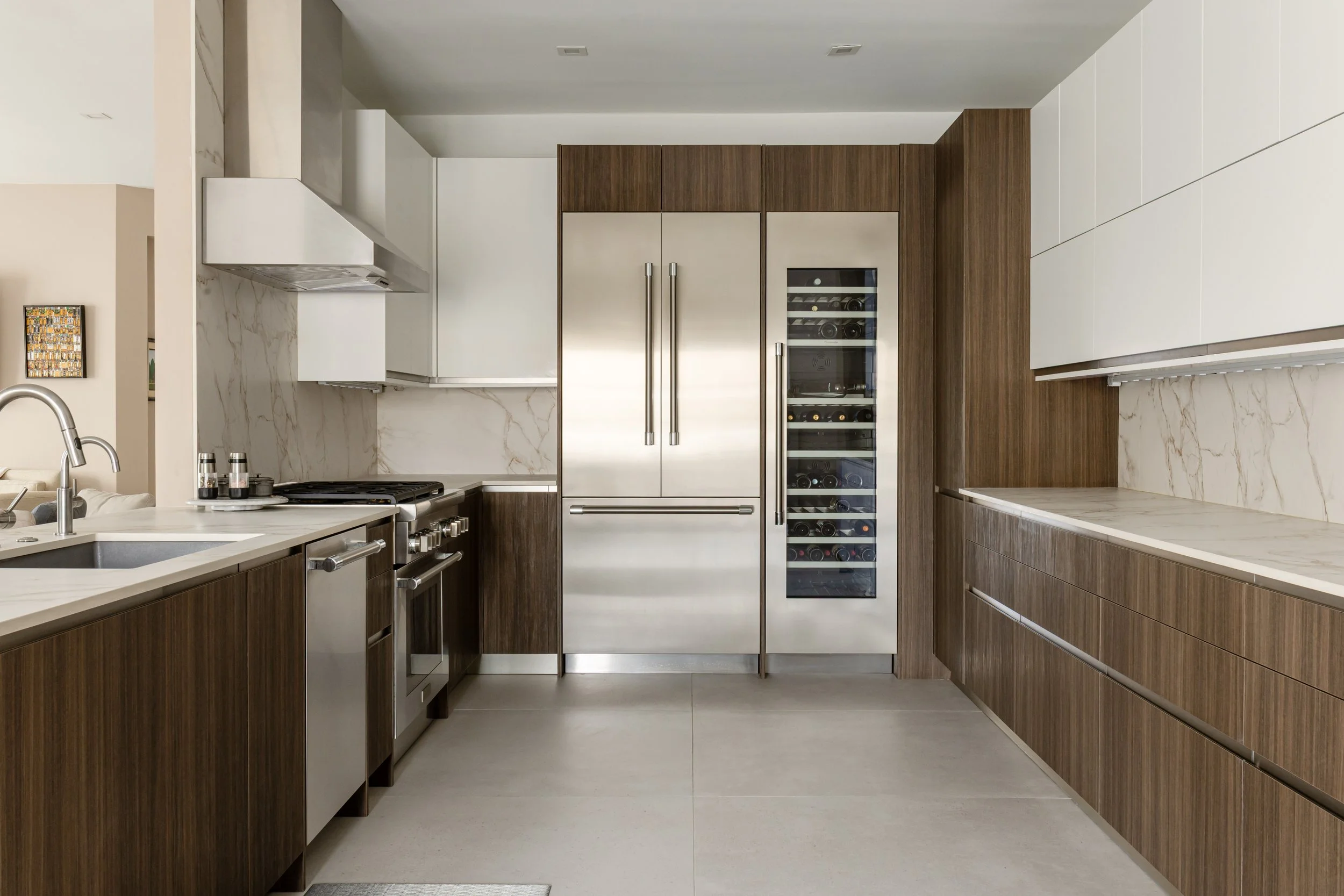 Modern kitchen with white marble countertops, wooden cabinets, and stainless steel appliances including a double-door refrigerator and wine cooler.
