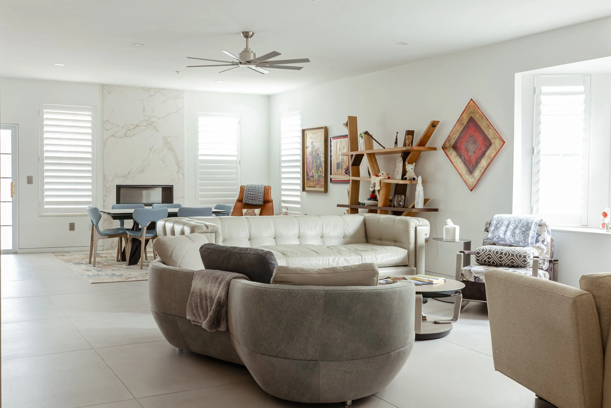 Living room with white sofa, gray chair with blanket, armchair near window, painting on wall, modern shelving unit with decorations, dining table with chairs, ceiling fan, bright natural light.