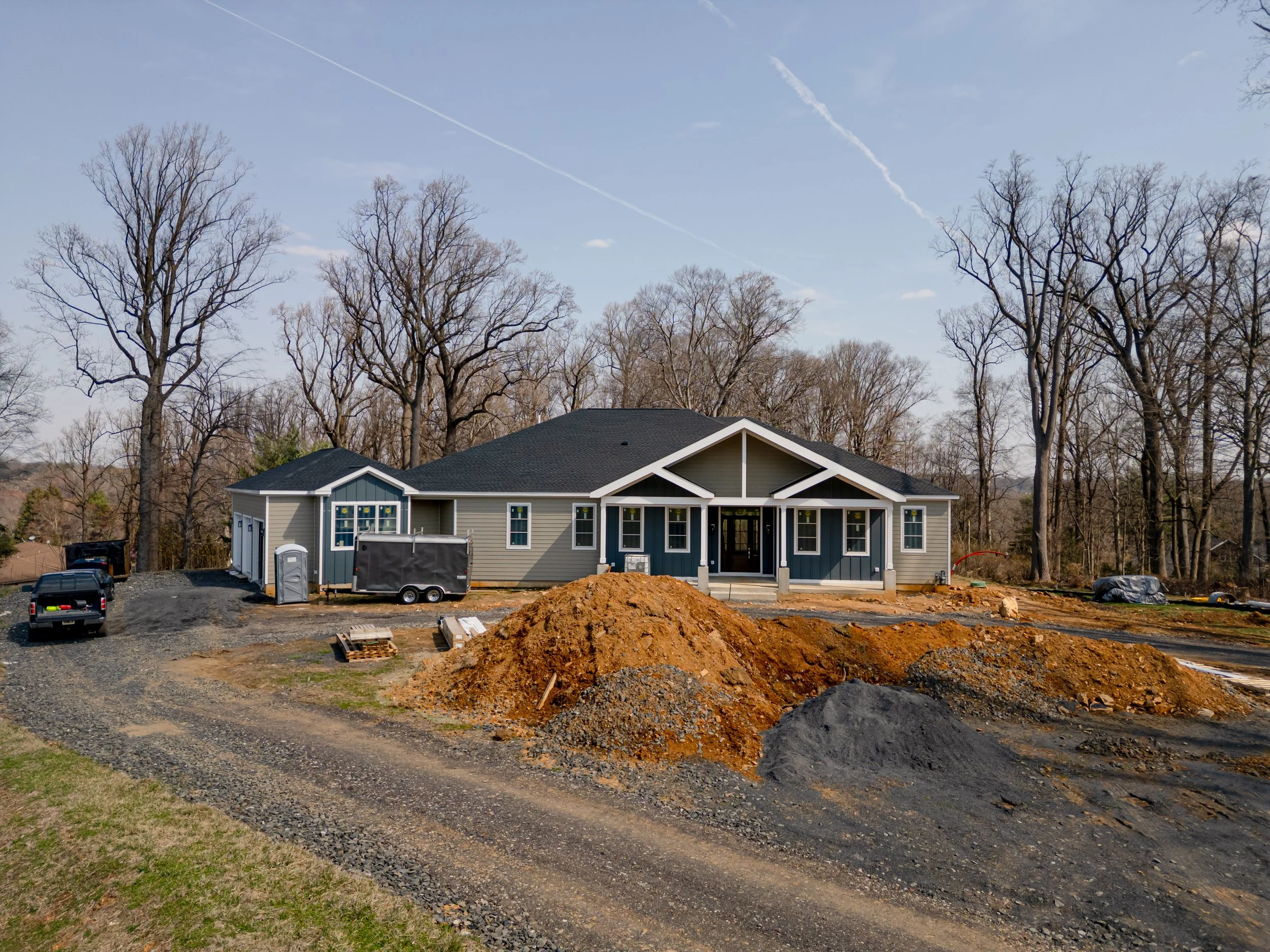 Under construction house with piles of dirt and construction materials in front, trees in background, and a clear sky.