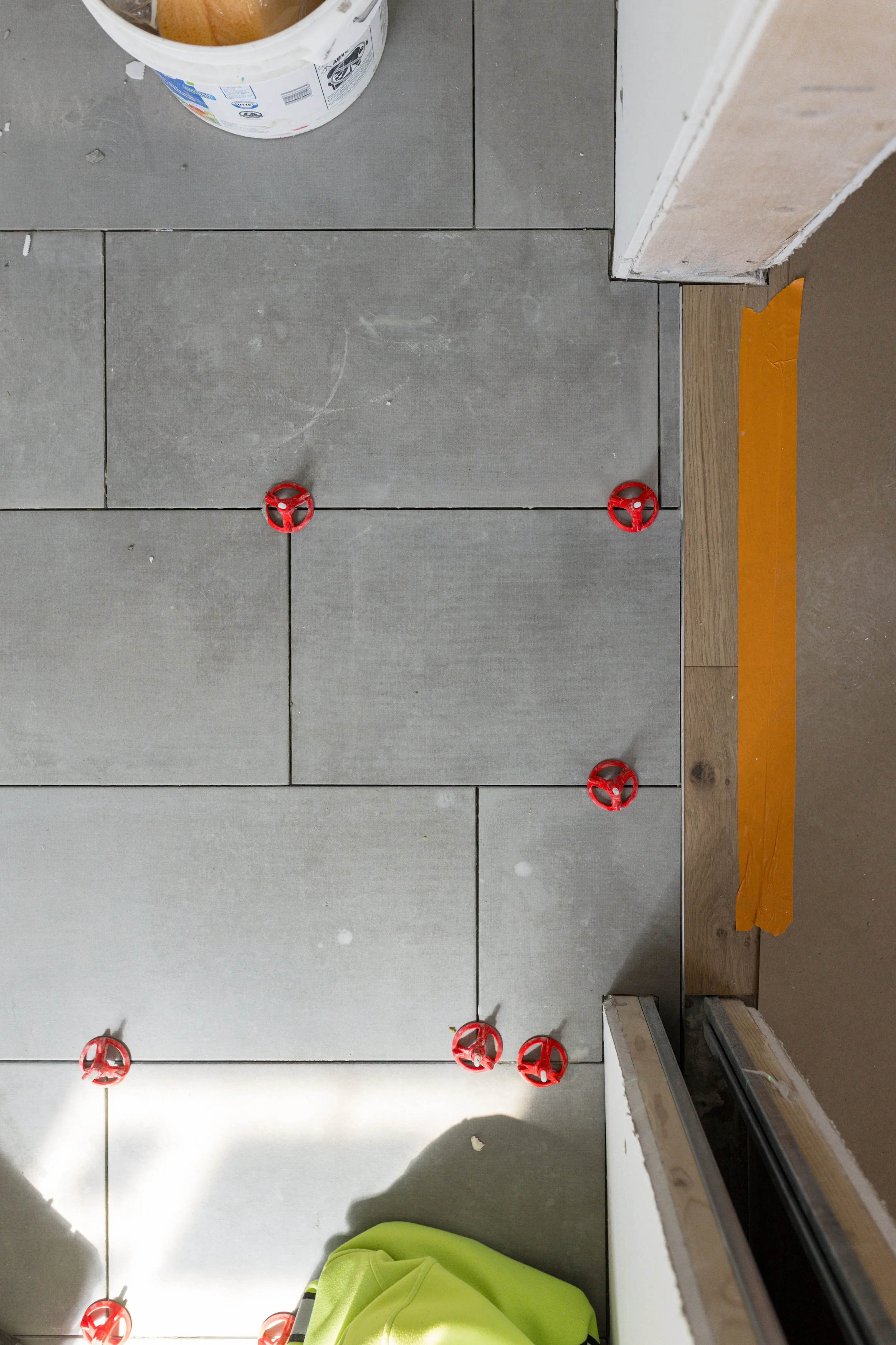 New tile floor being installed with red spacers in place to align tiles, and construction tools and materials visible in the surroundings.