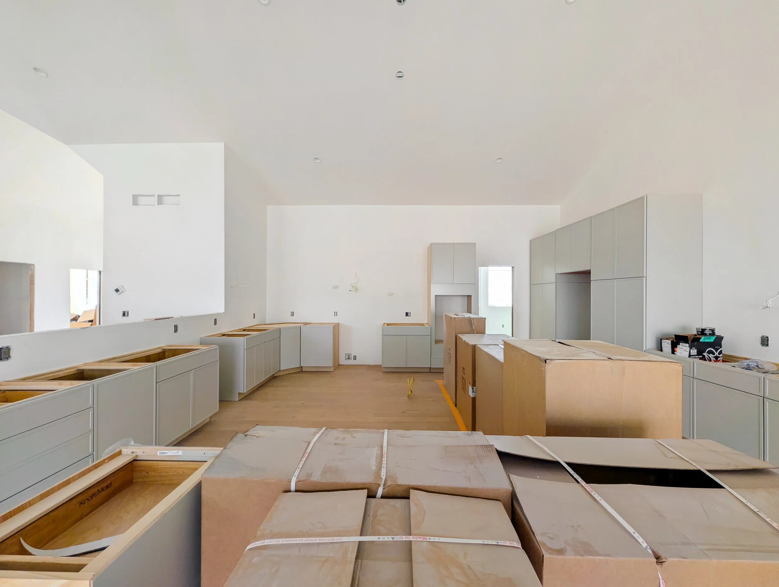 Construction site of a modern kitchen with white walls and cabinetry, some cabinets are unfinished, with cardboard boxes and packaging materials on the floor.