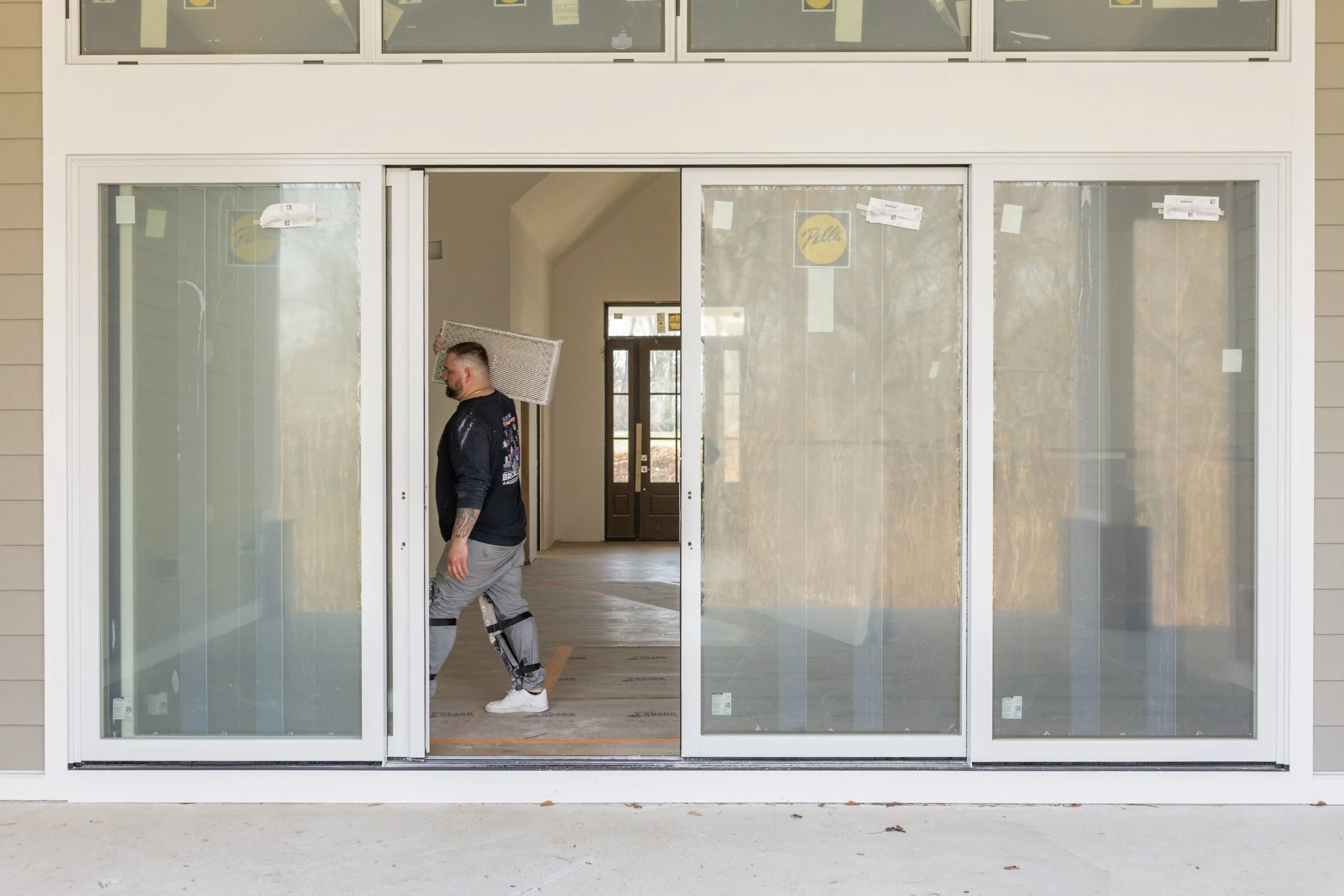 A man carrying a box in a house under construction, seen through a large sliding glass door. The house has an open floor plan with a front door visible in the background.