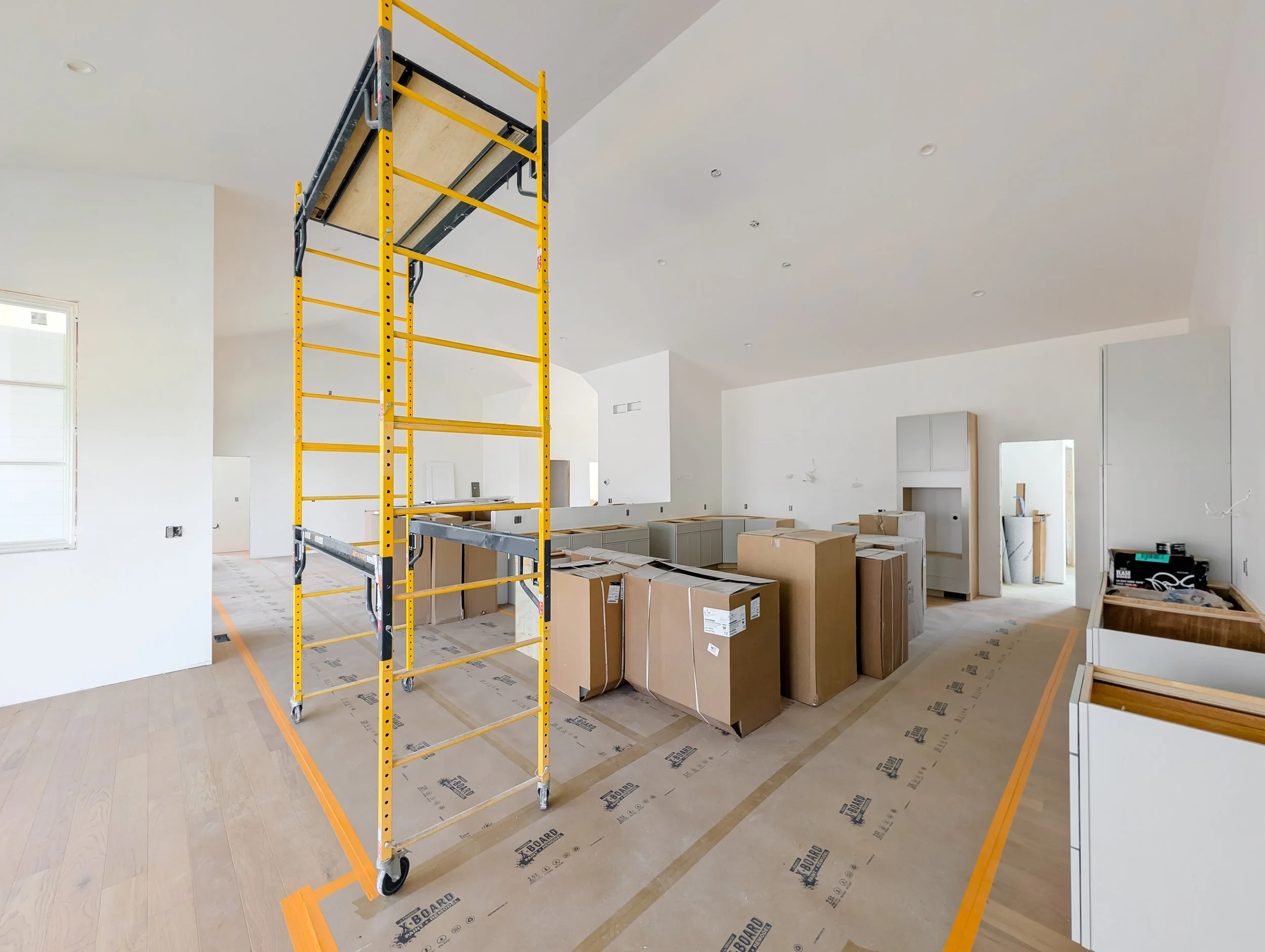 Empty room under construction with a yellow scaffolding, numerous packed boxes, and ongoing cabinetry installation, with white walls and wood flooring.
