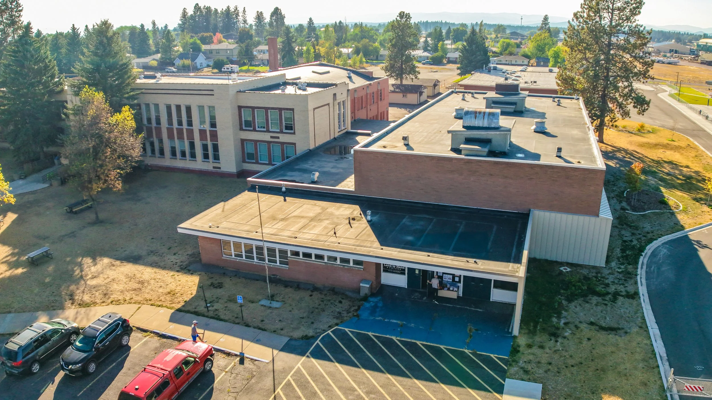 An aerial view of a school building with a main entrance, parking lot filled with cars, and surrounding trees in a suburban area.
