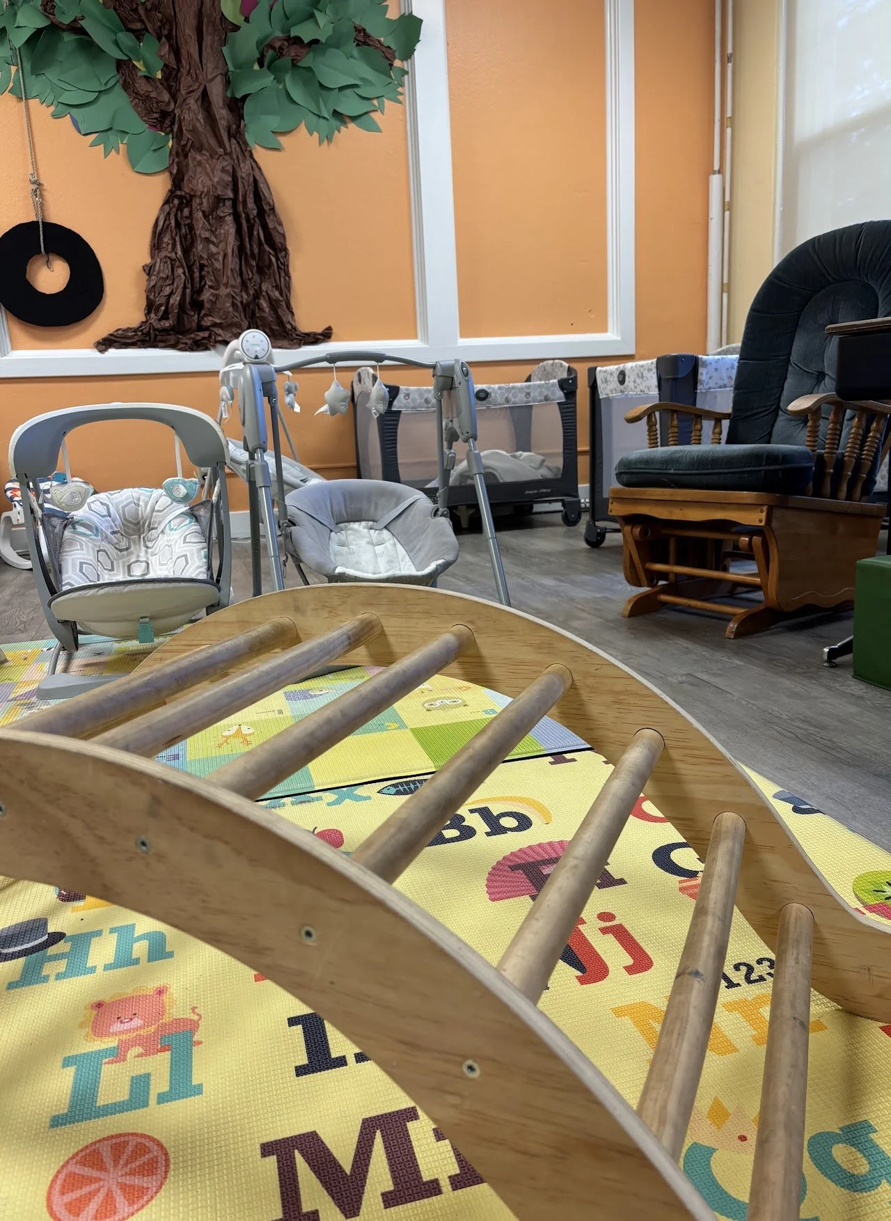 A playroom with a large orange wall decorated with a paper tree and a black tire swing. The room contains baby cribs, car seats, and a wooden rocking chair, with a colorful alphabet foam mat on the floor.
