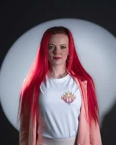 Vrouw met lang rood haar die naar de camera kijkt, wit T-shirt met een kleurrijk logo, donkere achtergrond met een lichtbol.