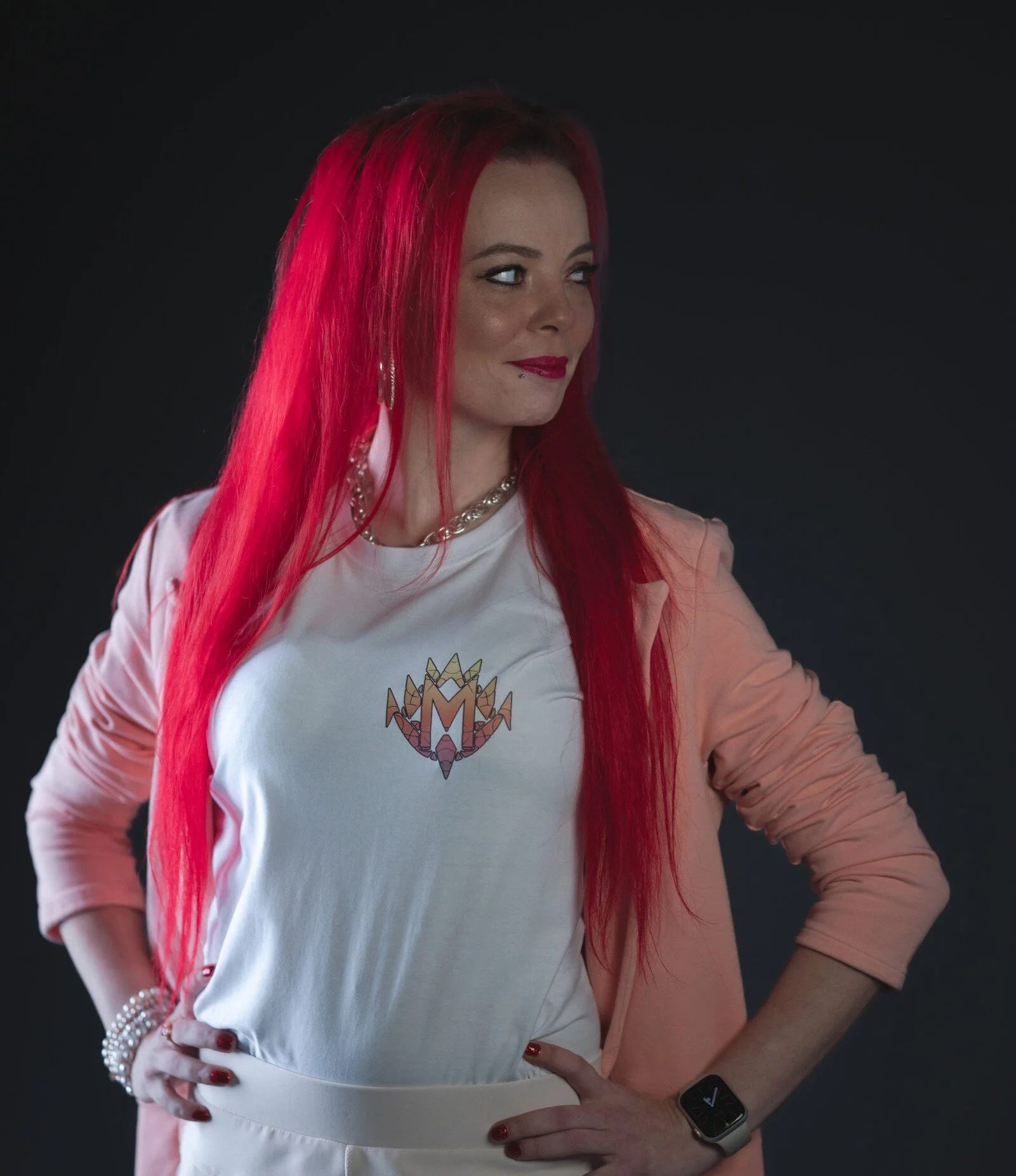 Portret van een vrouw met rood haar, gewapend met een witte T-shirt met een logo, sieraden en een smartwatch, poseert tegen een zwarte achtergrond.