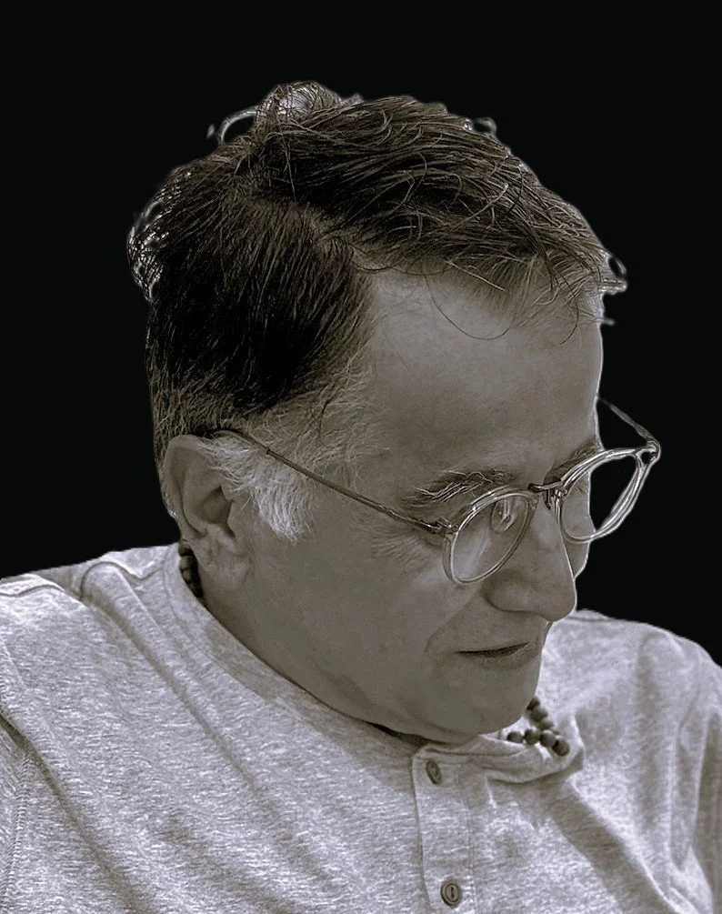 A black-and-white portrait of an older man with lenses and short, dark hair. He is wearing a light-colored shirt and a beaded necklace, looking downward with a serious expression.