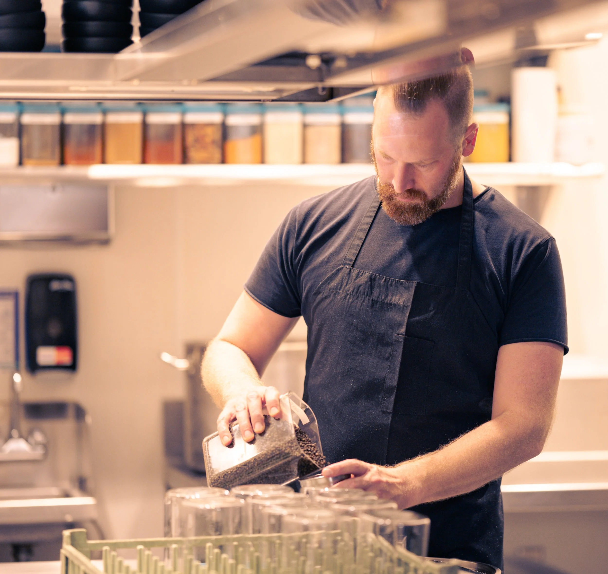 A chef in a black T-shirt and apron pouring black sesame seeds into a container in a kitchen.