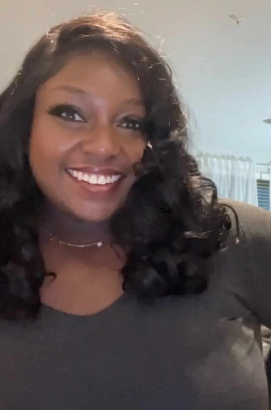 A woman with dark hair styled in loose curls, smiling, wearing makeup, a choker necklace, and a dark top, sitting indoors with a background that includes a window with curtains.