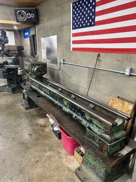 Long, old green metal lathe machine in a workshop with American flag on wall.