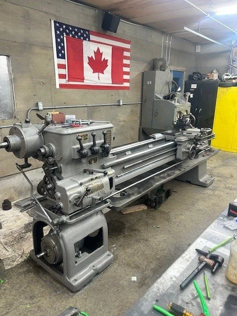 Metal lathe machine in a workshop with a Canadian and an American flag hanging on the wall.