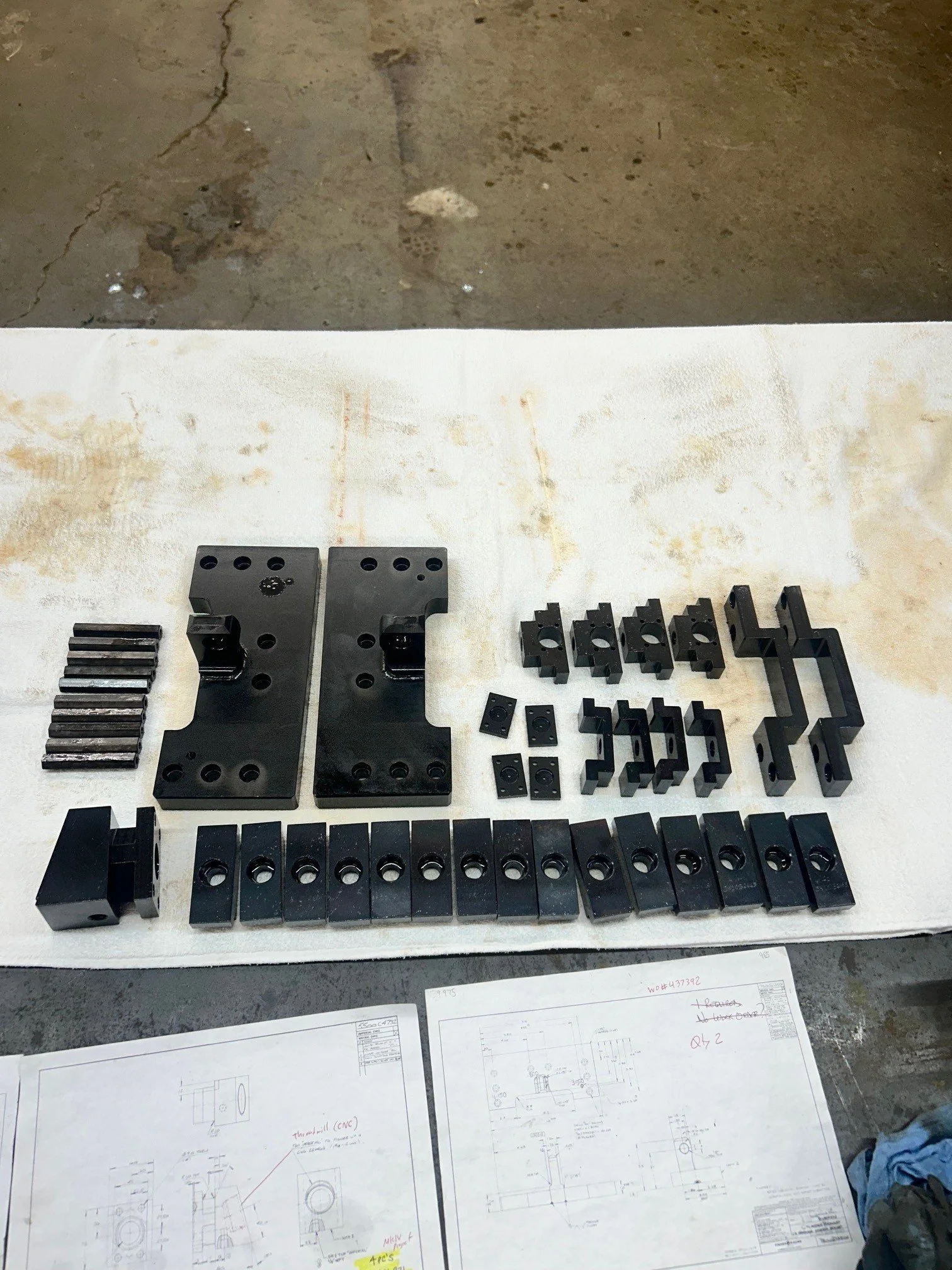 Black metal mechanical parts and components arranged on a white table with engineering drawings beneath. The parts include blocks, plates, and brackets of various shapes and sizes.