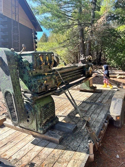 A large, old, green industrial machine with peeling paint and rust is mounted on a flatbed trailer in a wooded area with tall trees. A young girl in a gray hoodie, yellow rain boots, and pink pants is nearby, handling a hose or a tool.