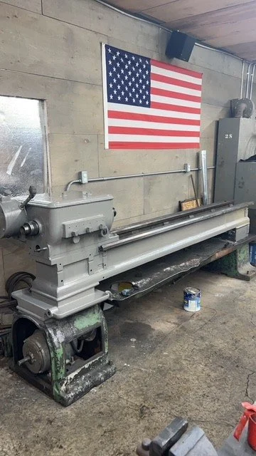 A large metal lathe machine in a workshop with an American flag hanging on the wall behind it.